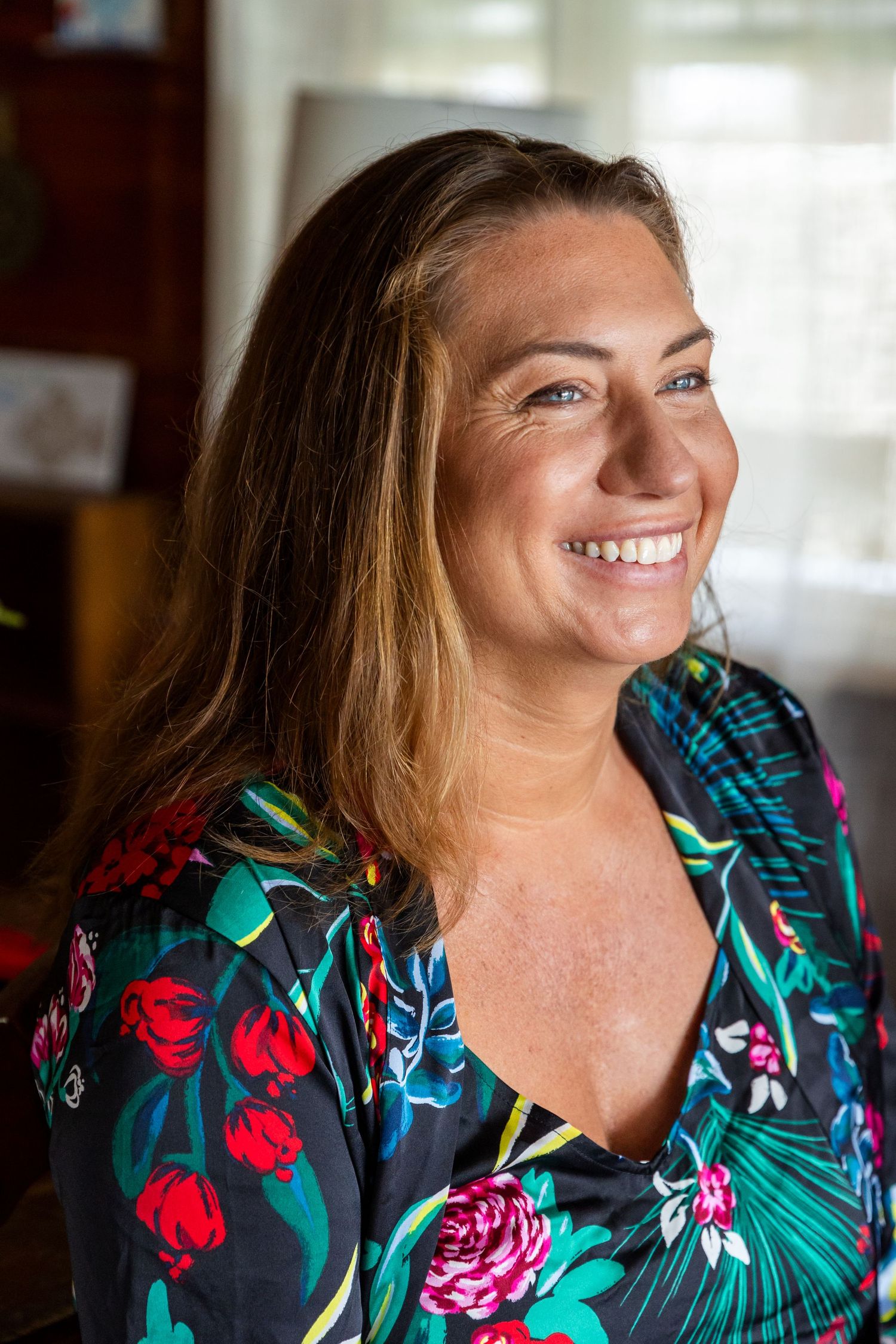 A person wearing a vibrant floral print dress smiles warmly in natural indoor lighting.
