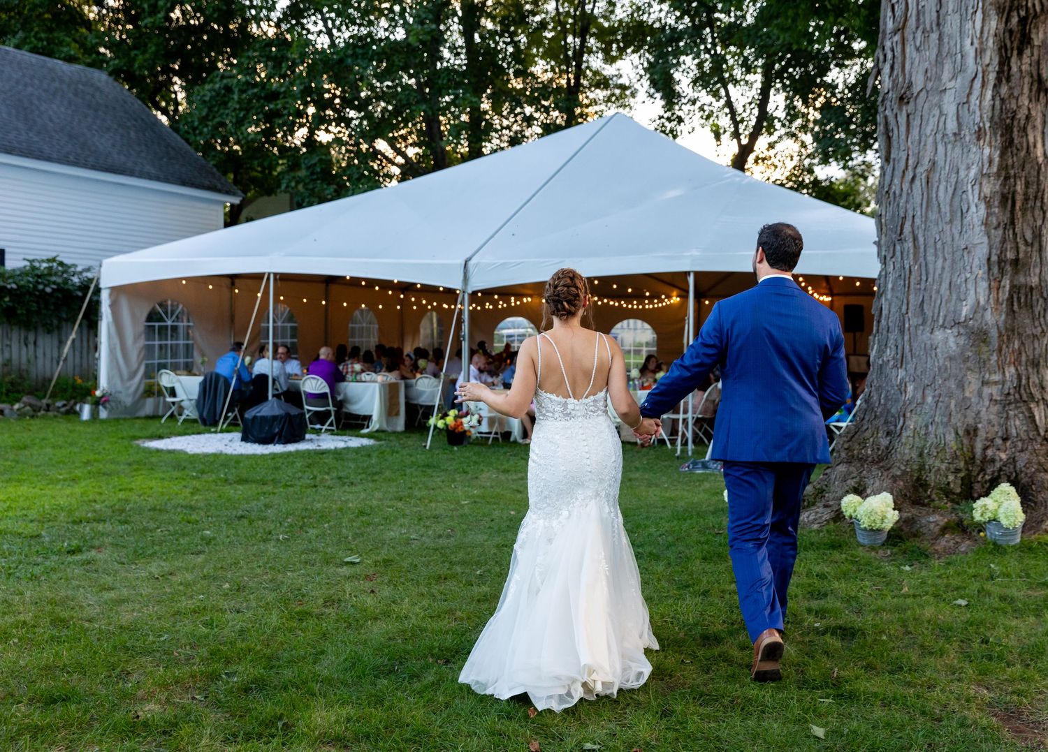 A couple in formal attire walking across a grass lawn with white tents and trees in the background at an outdoor event.