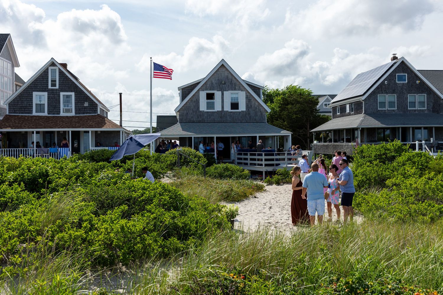 Beachfront vacation homes with an American flag and tourists enjoying a sandy path through coastal vegetation.