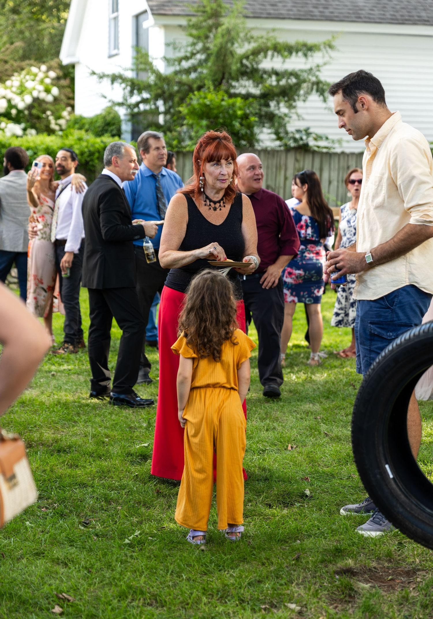 Guests gather at an outdoor party where a small child in a bright yellow outfit mingles with the crowd on a grassy lawn.