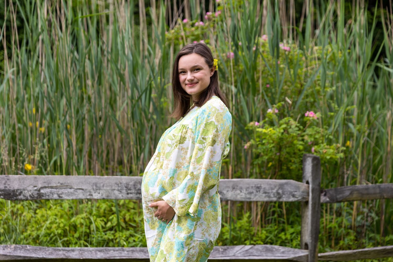 Expecting mother in a floral yellow and white dress poses by a rustic wooden fence with tall reeds in background.