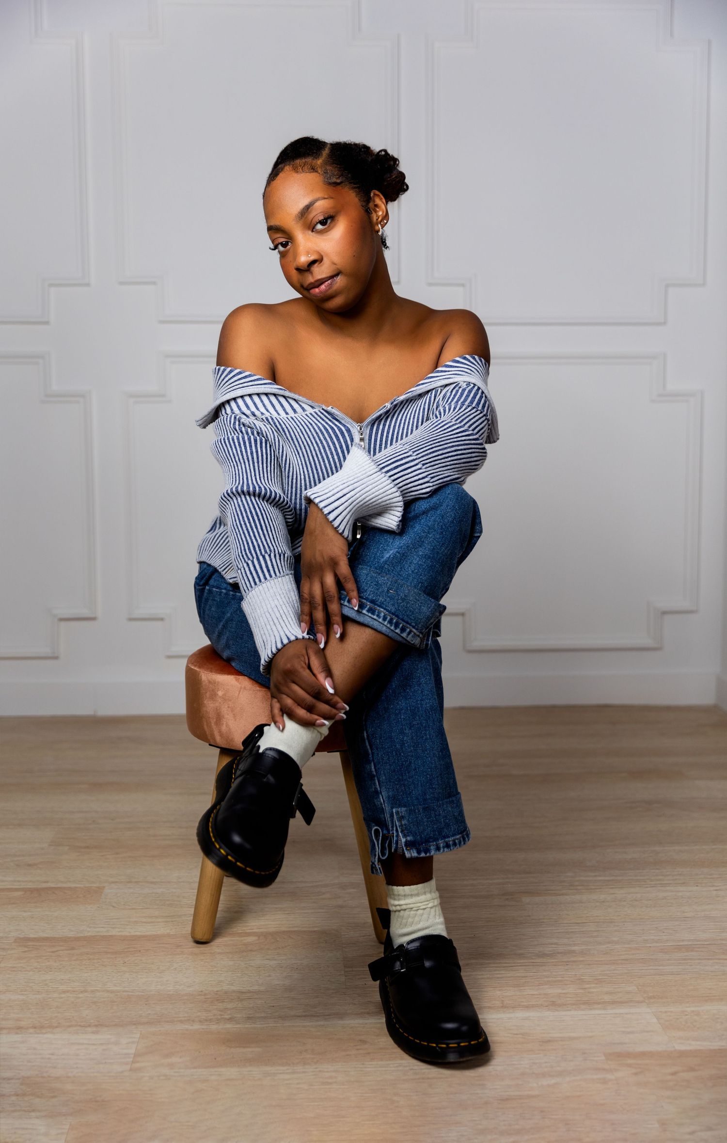 A person in casual denim and striped sweater poses confidently on wooden stool against white wall.