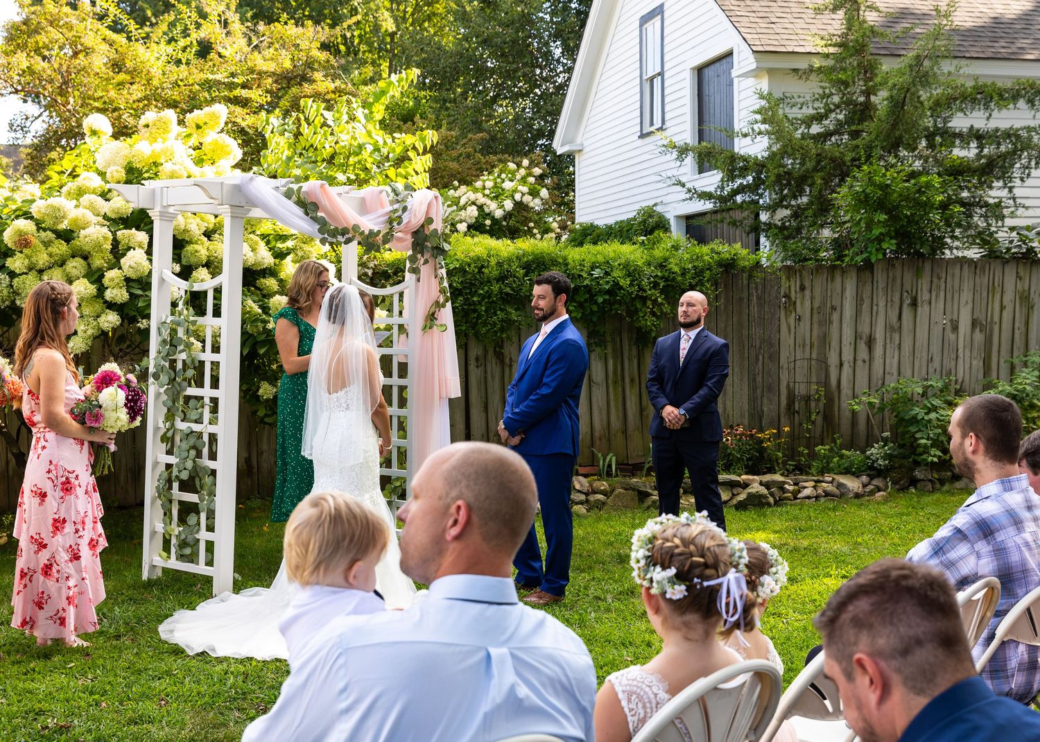 A garden wedding ceremony takes place on a sunny day with guests seated in white chairs and floral archway decorations.