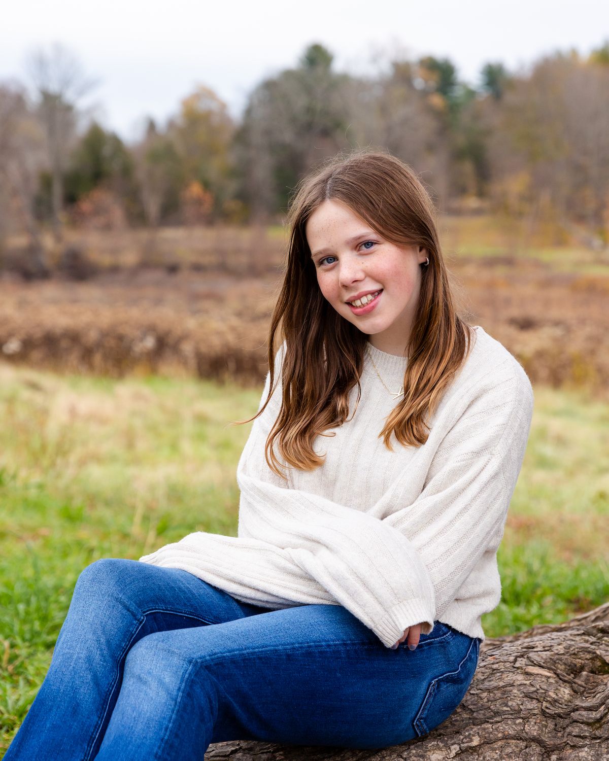 Someone in a cream sweater and jeans sits casually on a fallen tree trunk in an autumn field with trees in background.