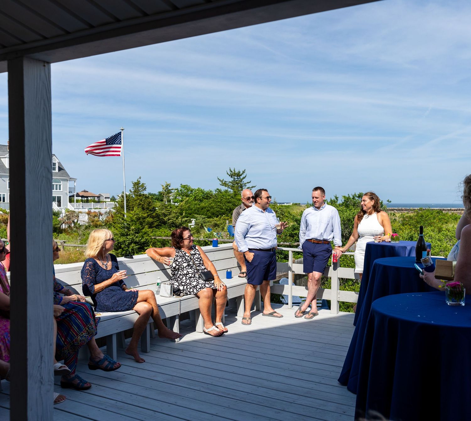 People gathered on a sunny deck overlooking the coast with an American flag flying in the blue sky above.