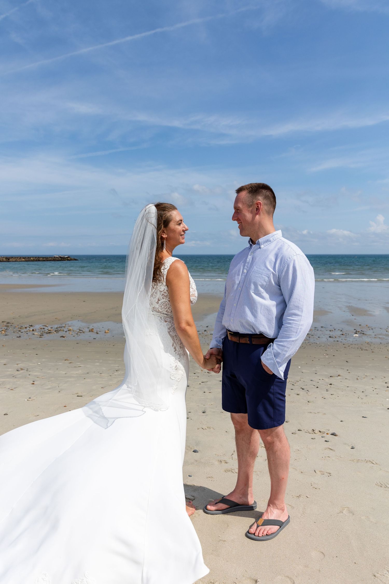 A couple on their wedding day stands together on a sunny beach with ocean waves and blue sky in the background.