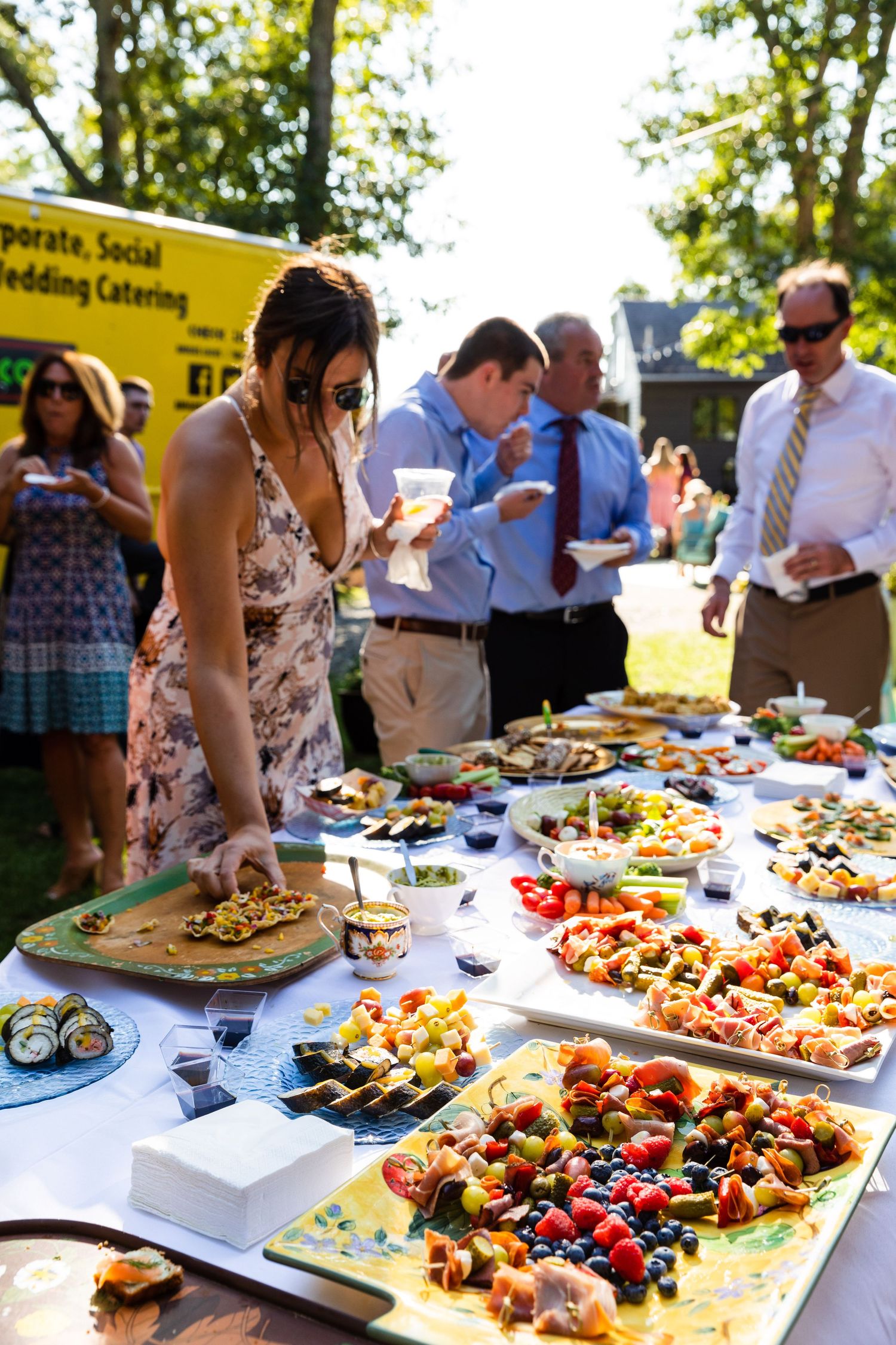 A buffet table filled with gourmet appetizers and party platters at an outdoor catering event in bright daylight.