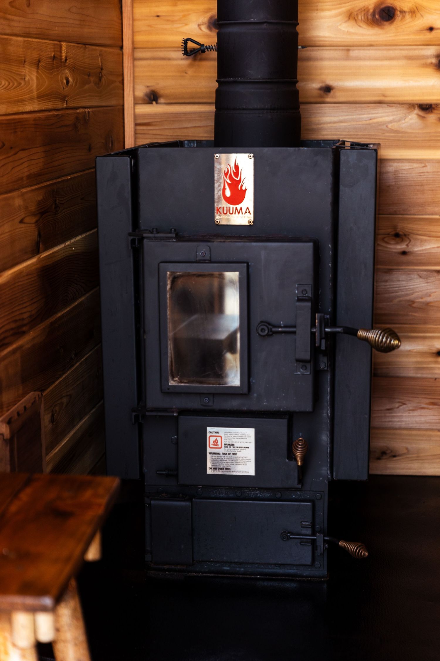 Black Kuuma wood burning stove with glass door installed against wooden walls in rustic cabin interior.