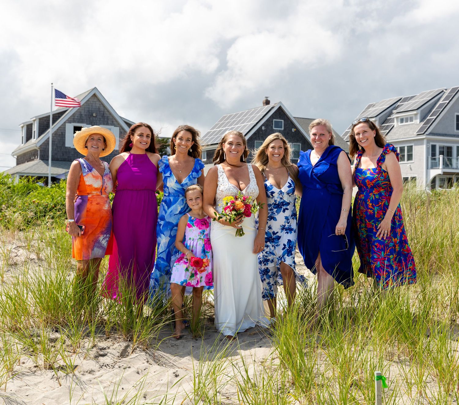 A group of friends in colorful summer dresses stand together on a sandy beach with coastal homes in the background.
