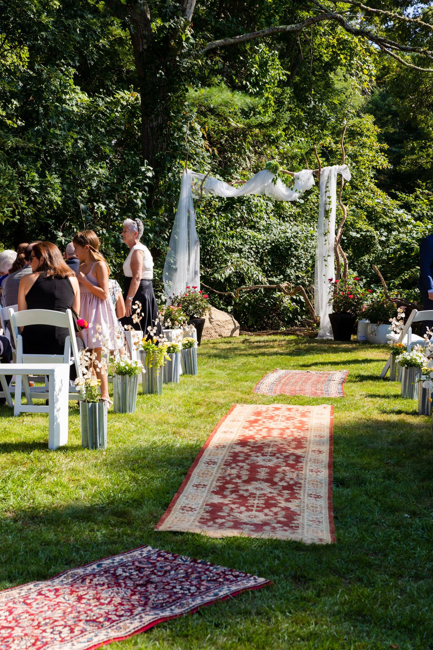 An outdoor wedding ceremony setup with vintage rugs leading to a white draped arch on a sunny lawn with white chairs.