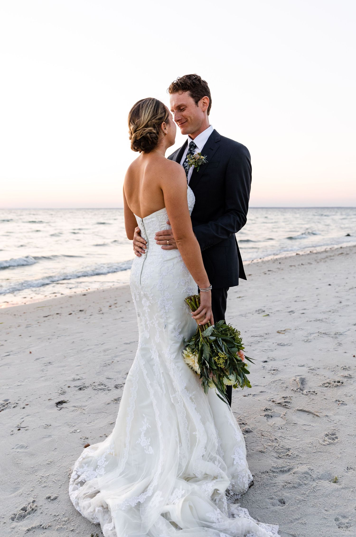 Newlyweds embrace on a sandy beach at sunset with bride in lace mermaid gown holding greenery bouquet.