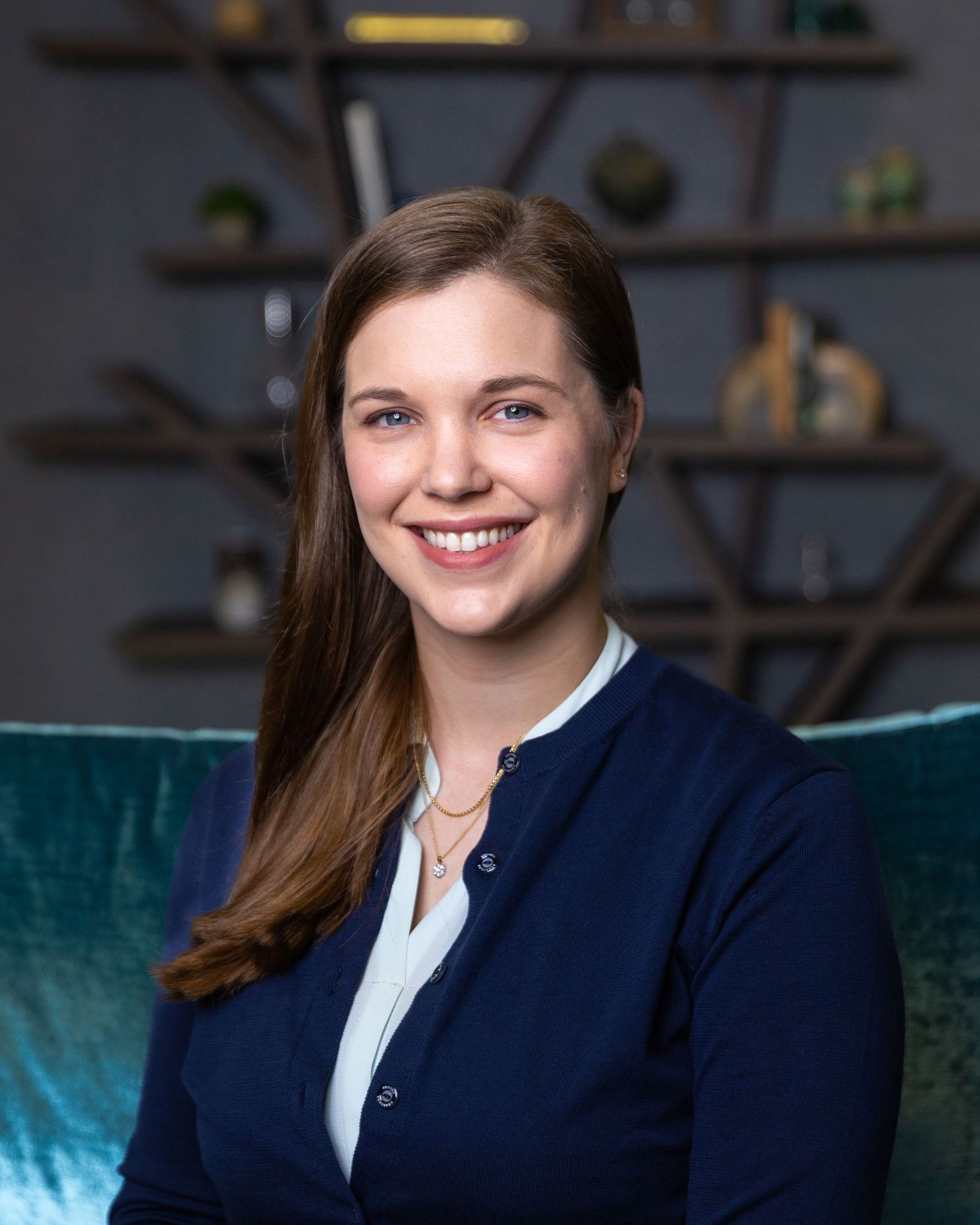 Professional headshot of a person in business attire seated on a teal velvet couch.
