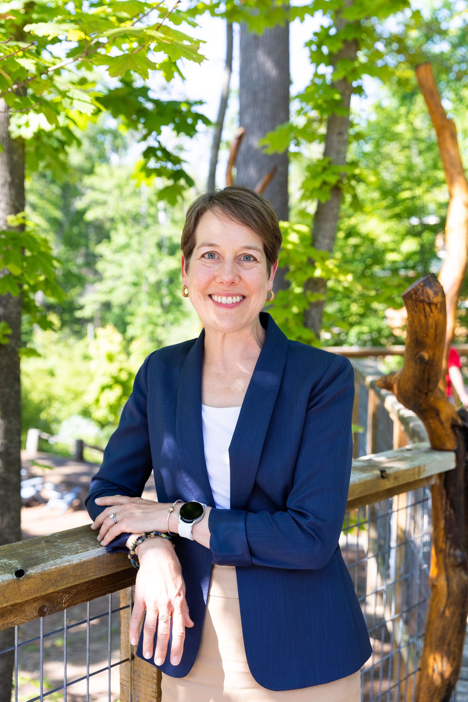 Professional outdoor portrait in navy blazer and white top standing by wooden railing with green forest background.