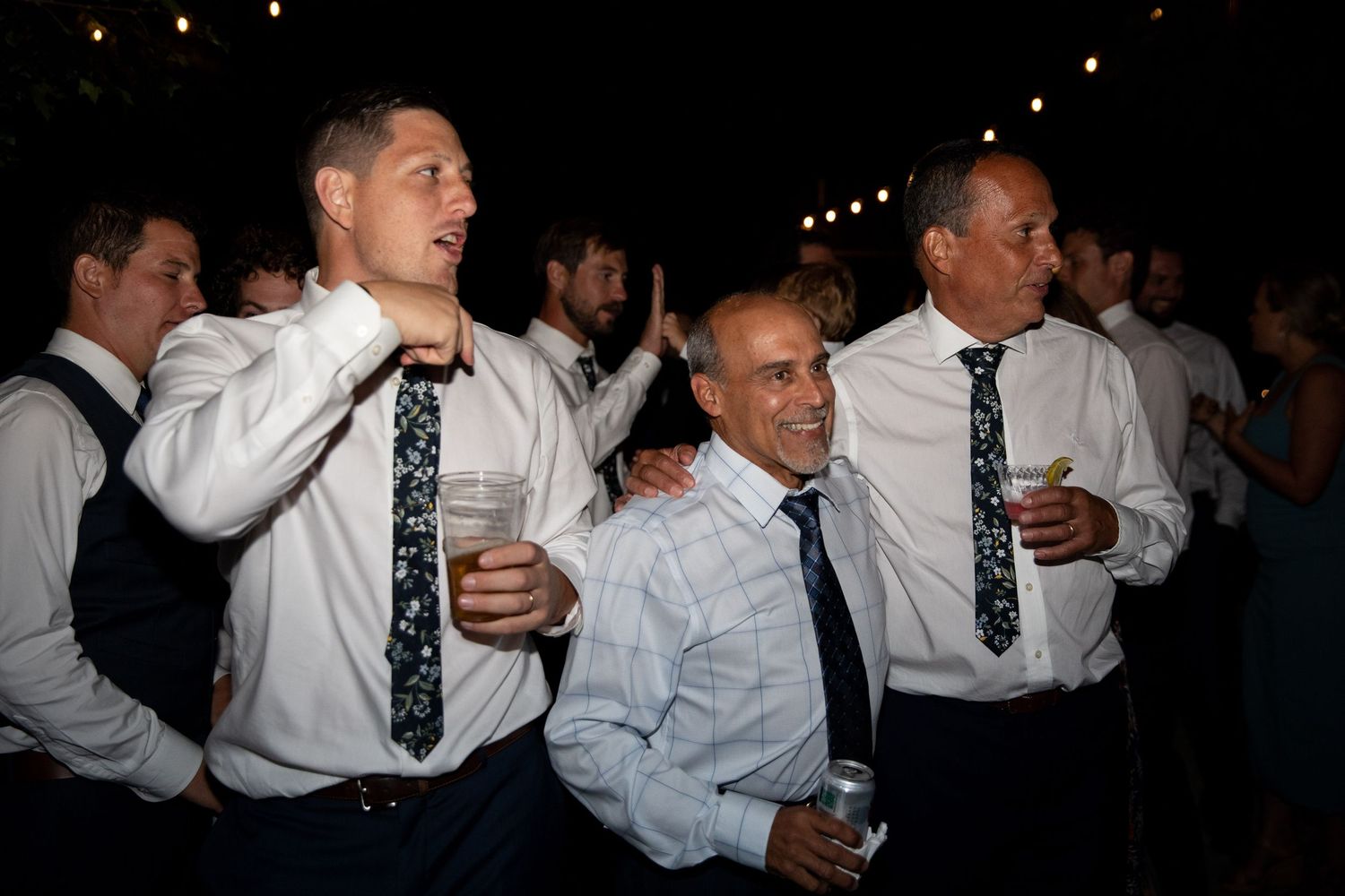Group of men in formal white shirts and ties celebrating at a nighttime event with drinks in hand.