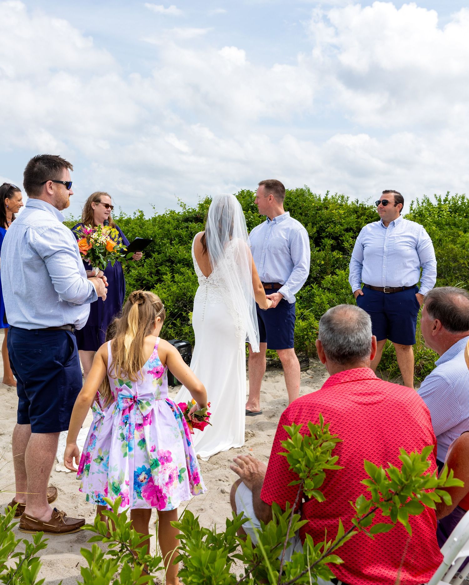 A beachside wedding ceremony with guests gathered on the sand under a bright blue sky with puffy white clouds.