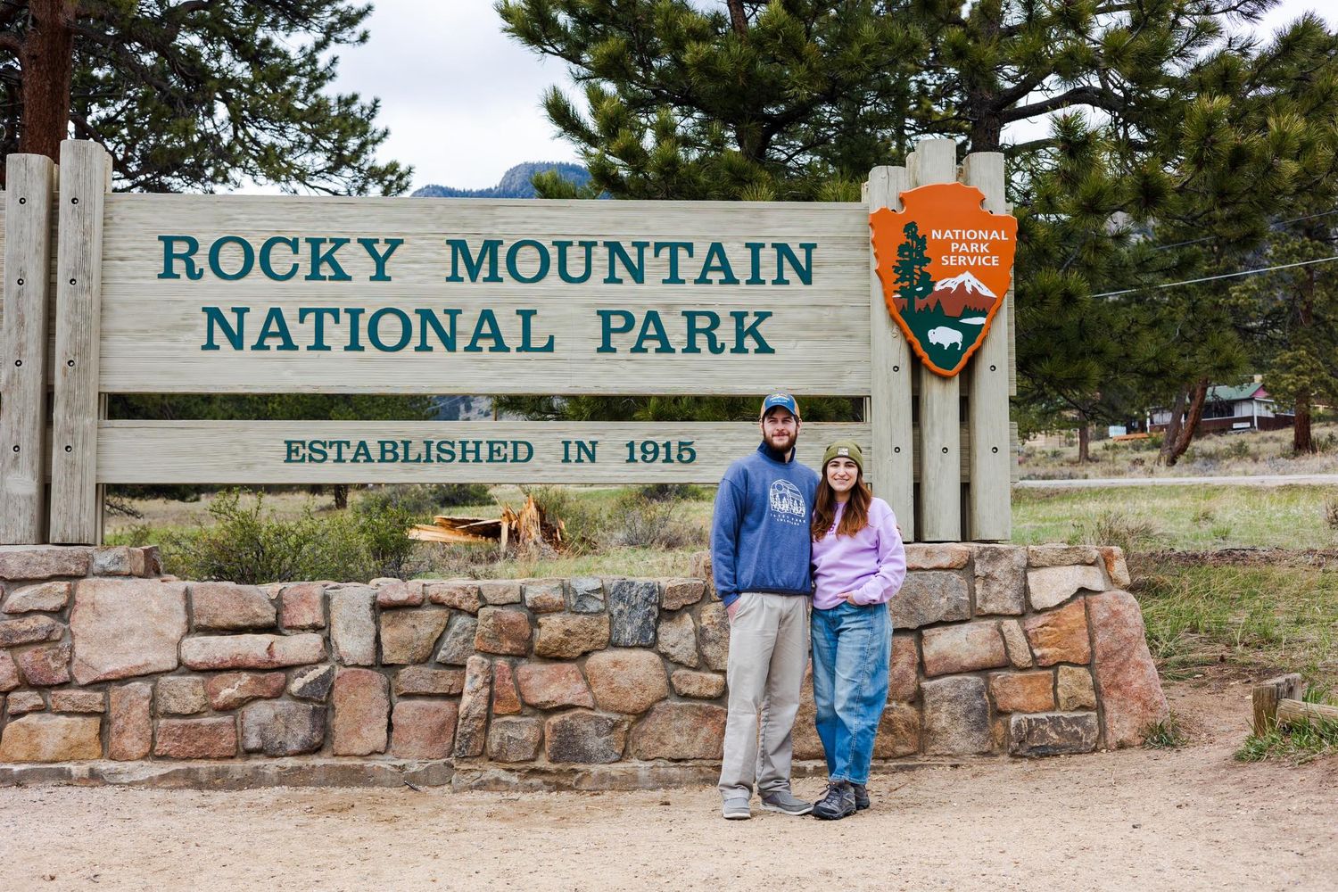 Two visitors pose next to the entrance sign of Rocky Mountain National Park established in 1915.