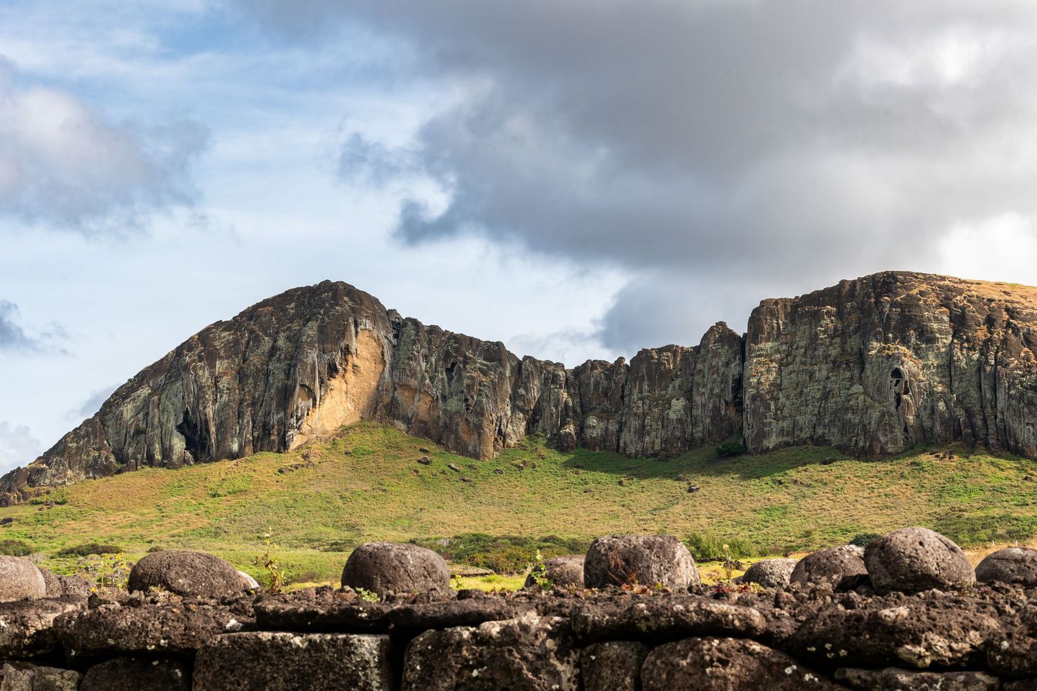 Dramatic rocky cliffs rise above a stone wall under moody clouds at Easter Island's ancient volcanic landscape.