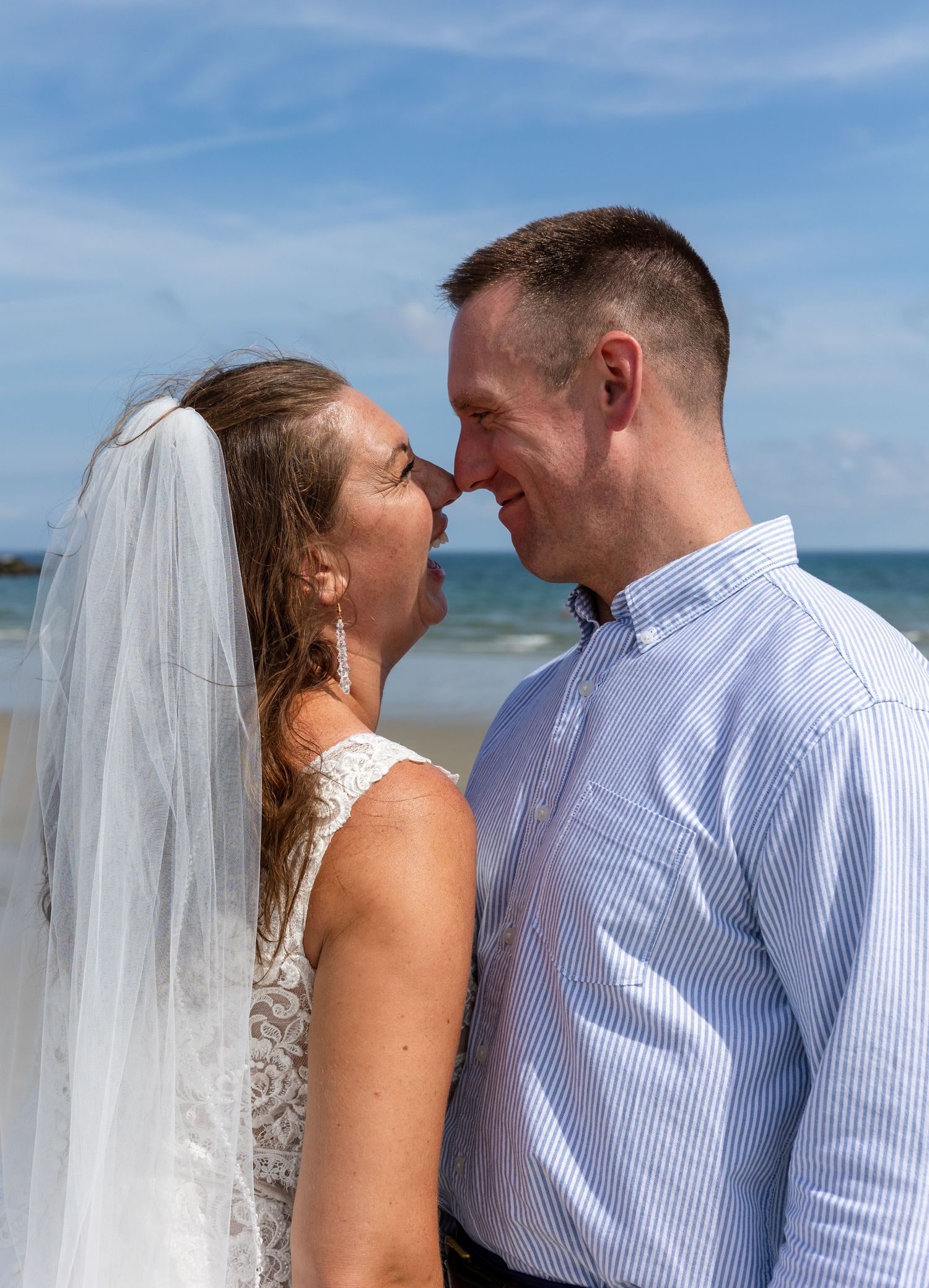 A couple sharing intimate moments on a sunny beach with ocean waves and blue sky in the background.