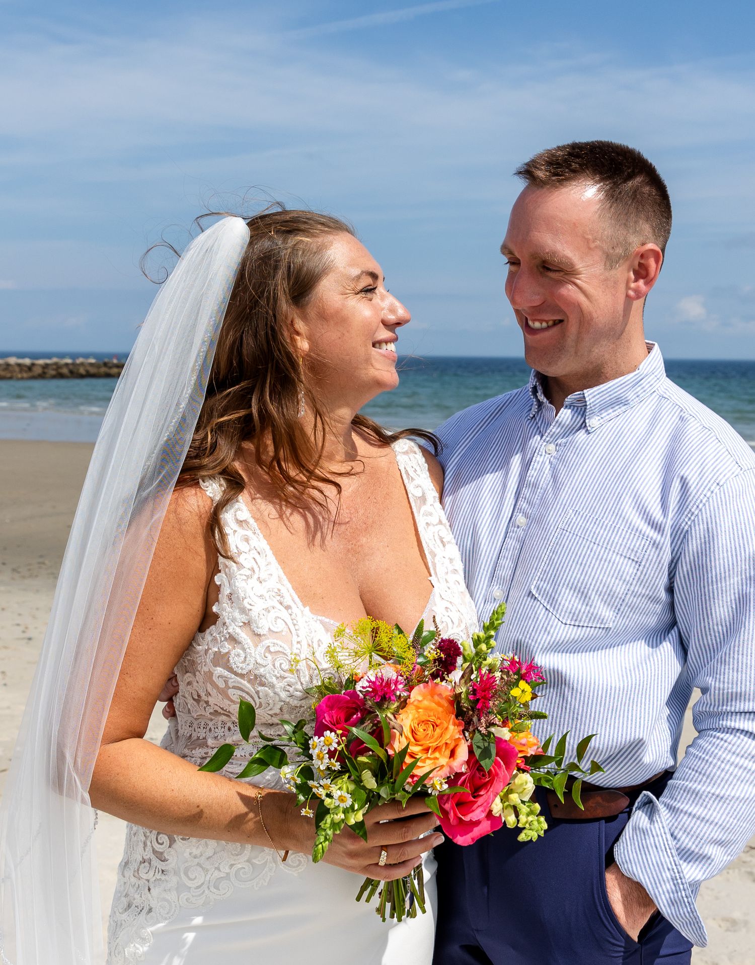 Couple shares joyful moment on beach with vibrant bouquet of mixed flowers on sunny day by ocean.