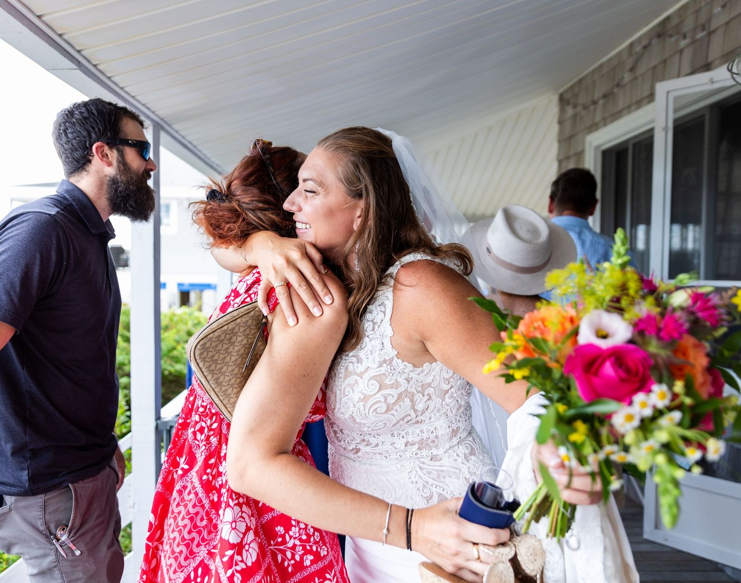 People embracing at an outdoor gathering with colorful floral arrangements and a porch in view.