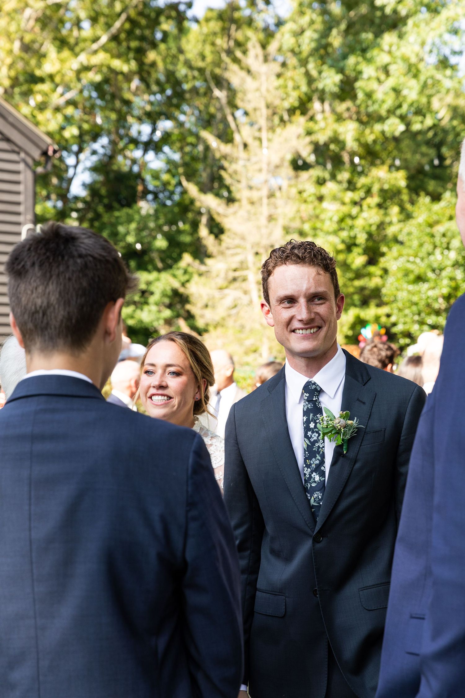 A group of people in navy suits stand together outdoors under trees on a bright sunny day.