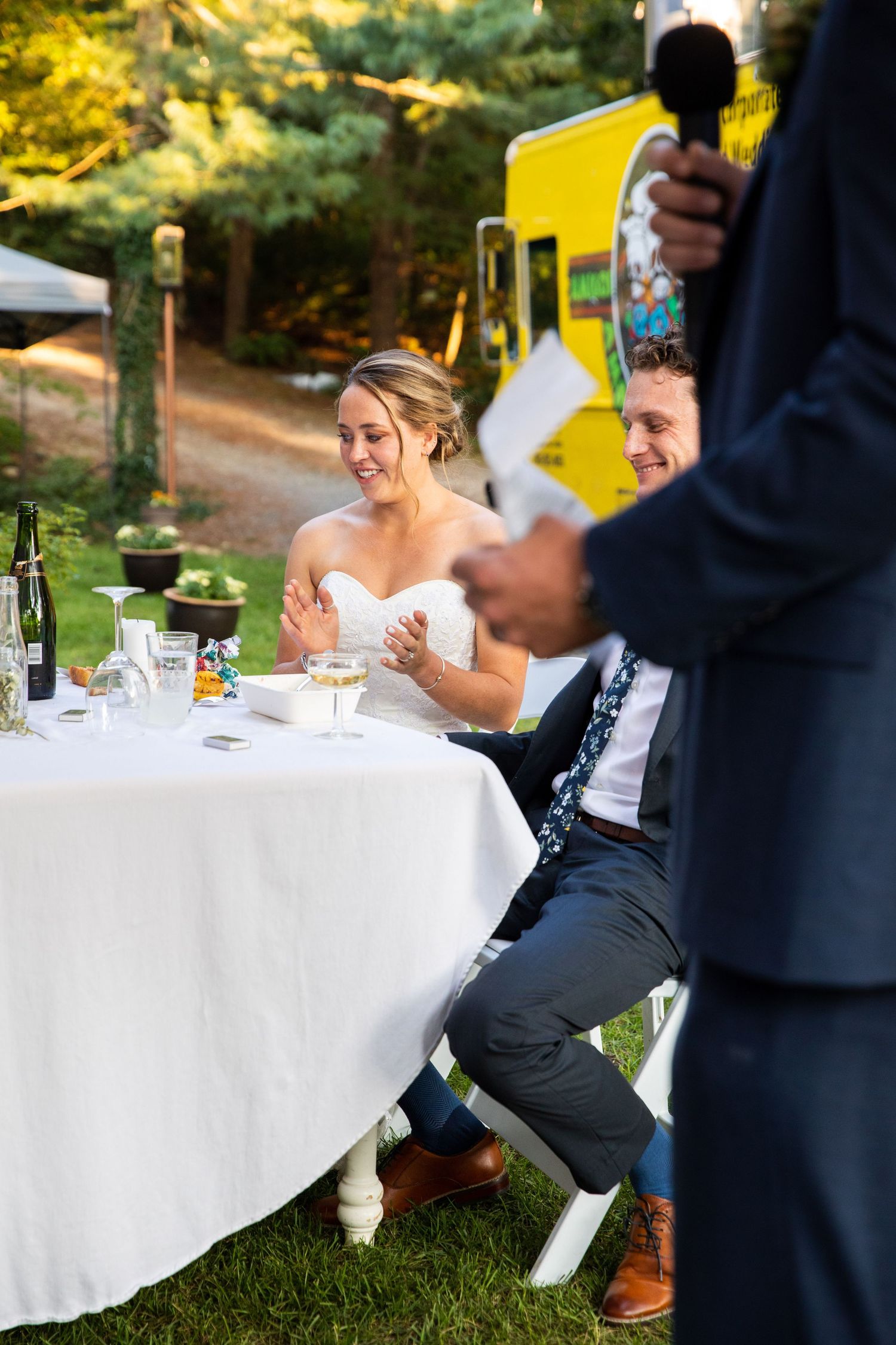 Wedding guests seated at an outdoor reception table with white linens as someone gives a speech during golden hour.