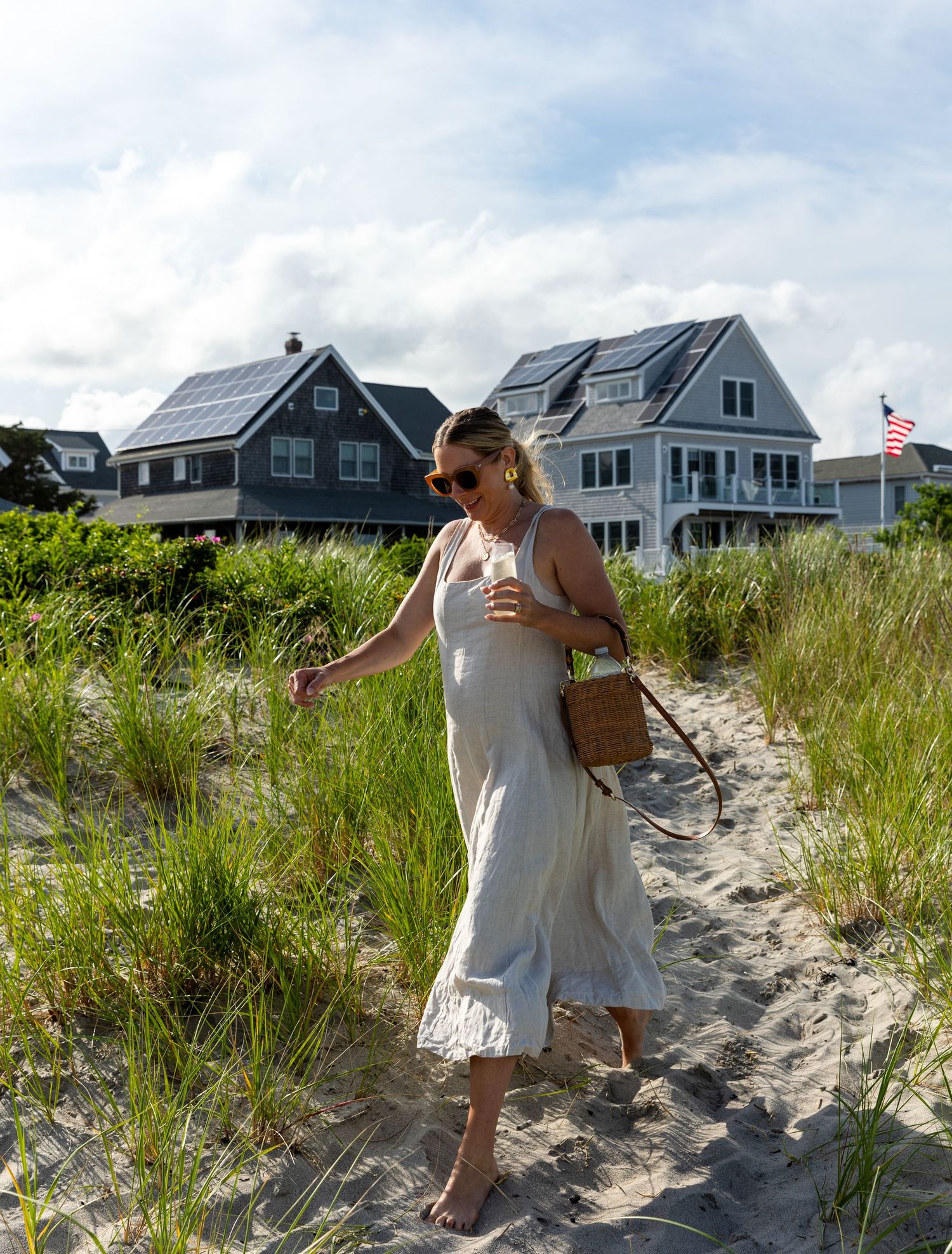 A person walks along a sandy beach path between coastal grasses with beachfront homes and solar panels in the background.