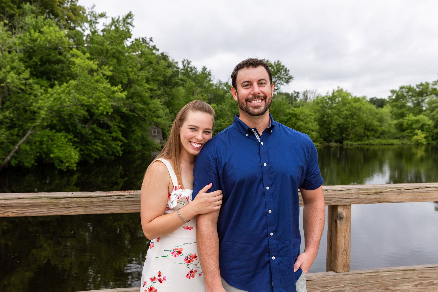 A couple stands together on a wooden dock overlooking a serene lake surrounded by green trees.