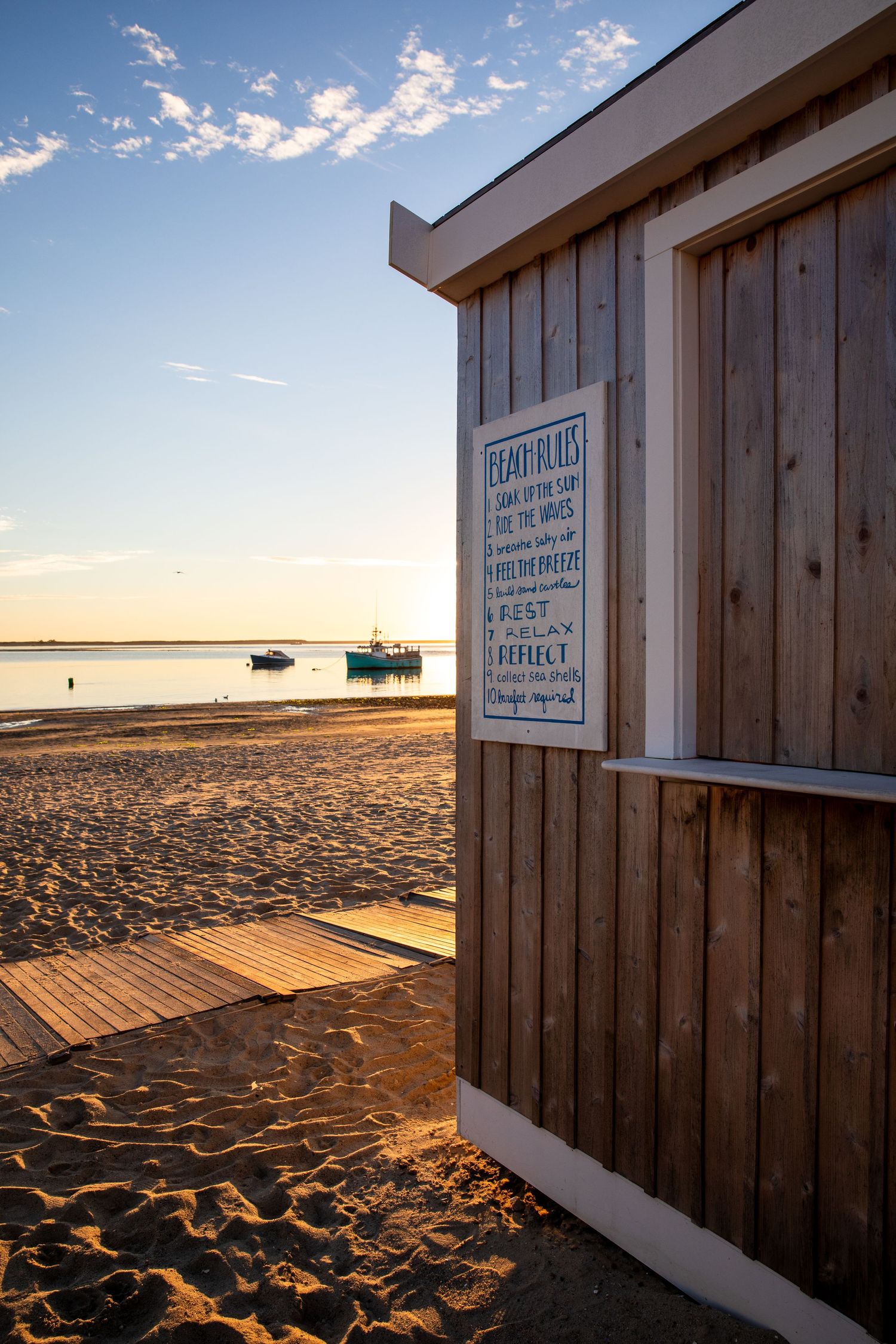 Wooden beach hut with notice board on sandy shore during sunset with boats in background.