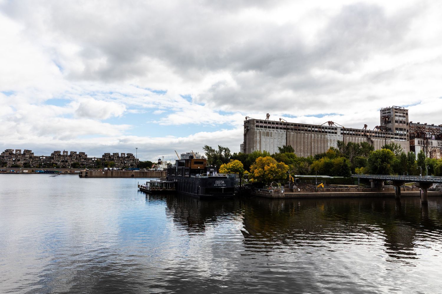 Industrial waterfront with large grain silos and buildings reflected in calm harbor waters.