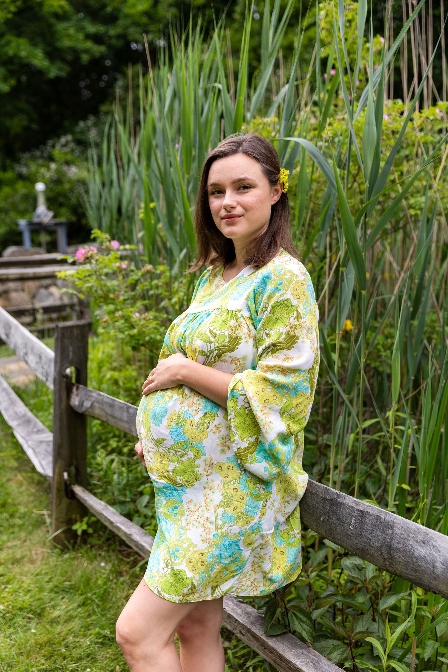 Expectant figure in floral yellow and green dress poses by rustic wooden fence near tall garden greenery.