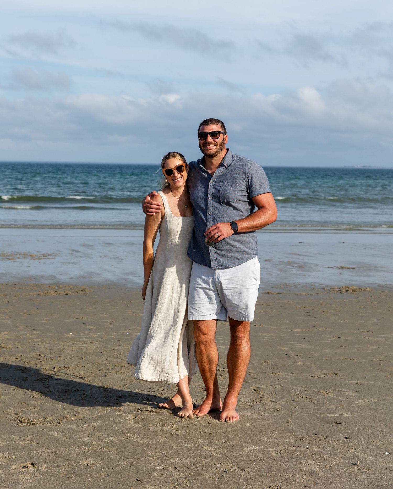 A couple standing together barefoot on a sandy beach with ocean waves and cloudy sky in the background.