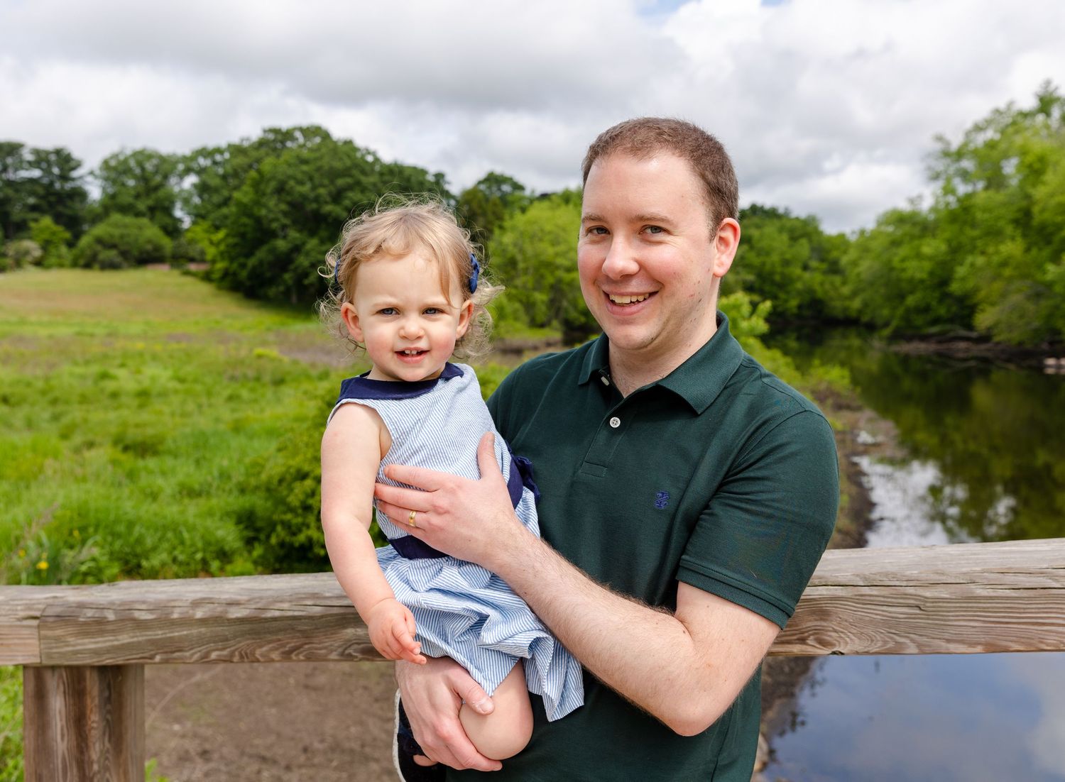 A parent and child stand together on a wooden bridge overlooking a tranquil river with green fields and trees in the background.