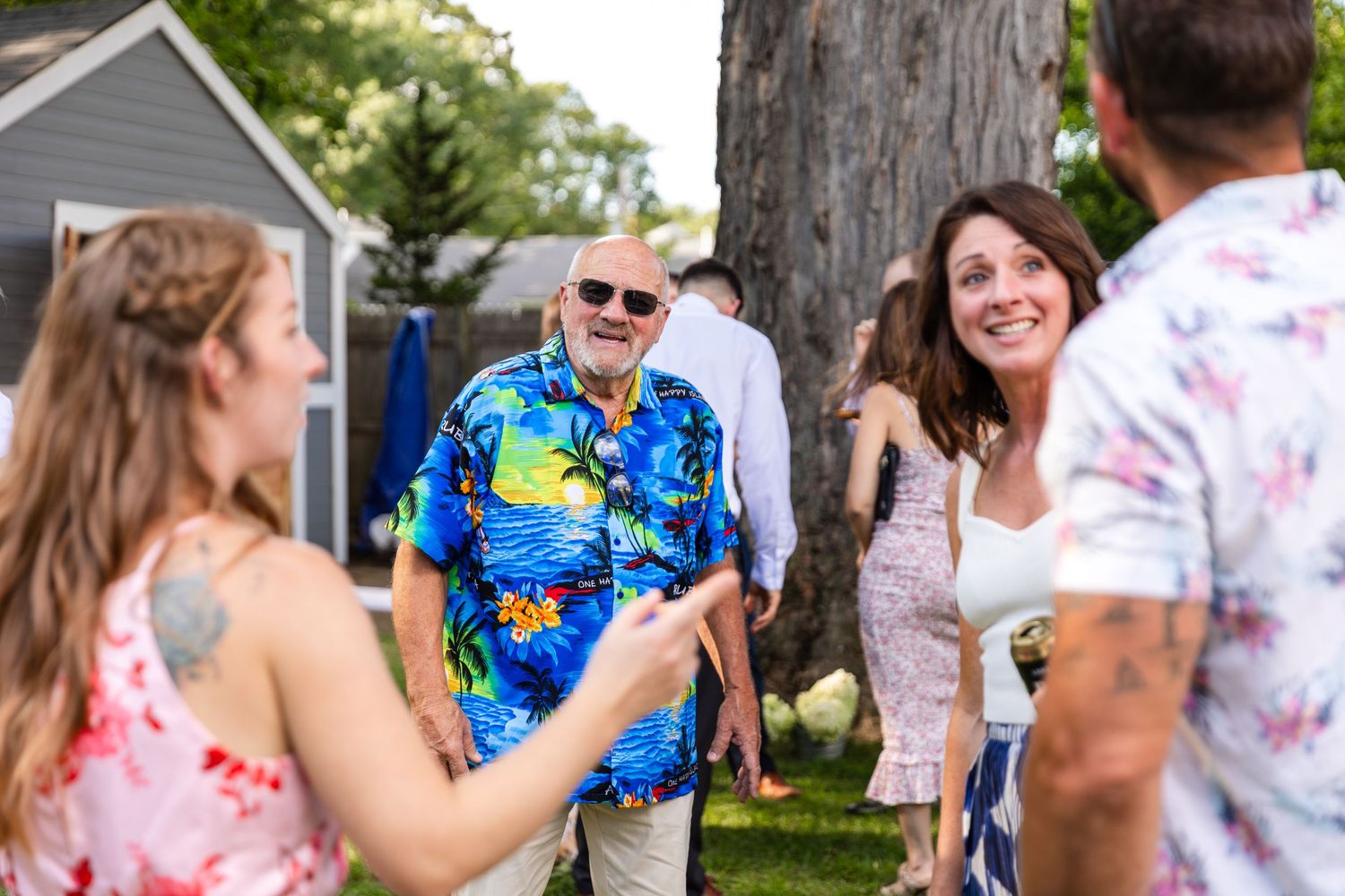 People at an outdoor summer gathering wearing colorful tie-dye and floral shirts enjoy drinks and conversation on a sunny day.