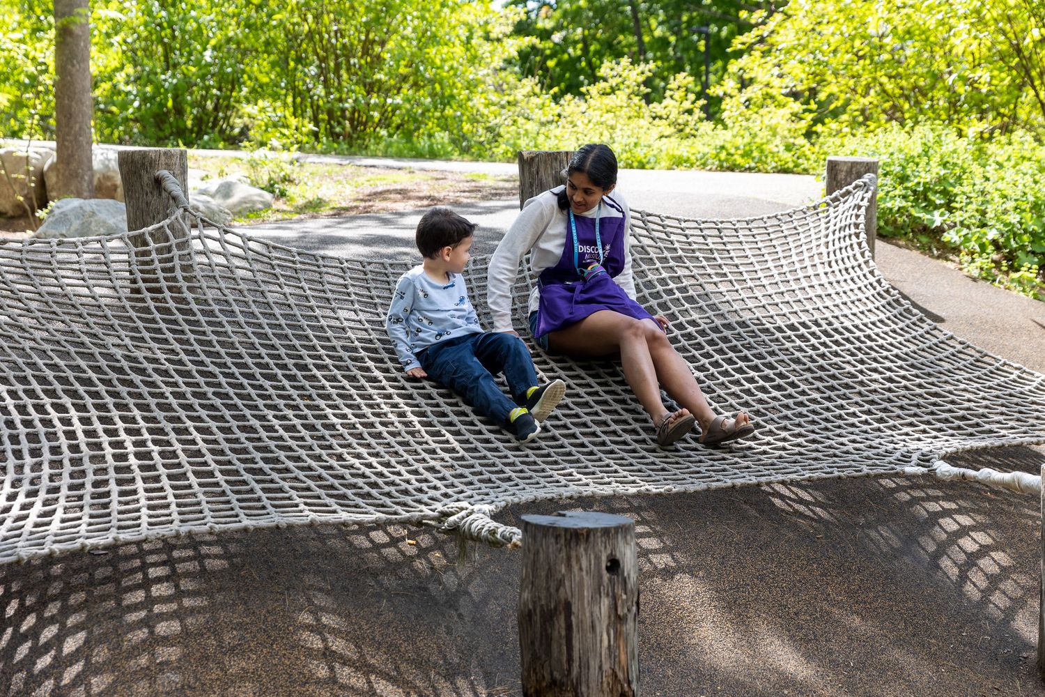 People relaxing together on a large outdoor rope hammock structure in a sunny garden setting.