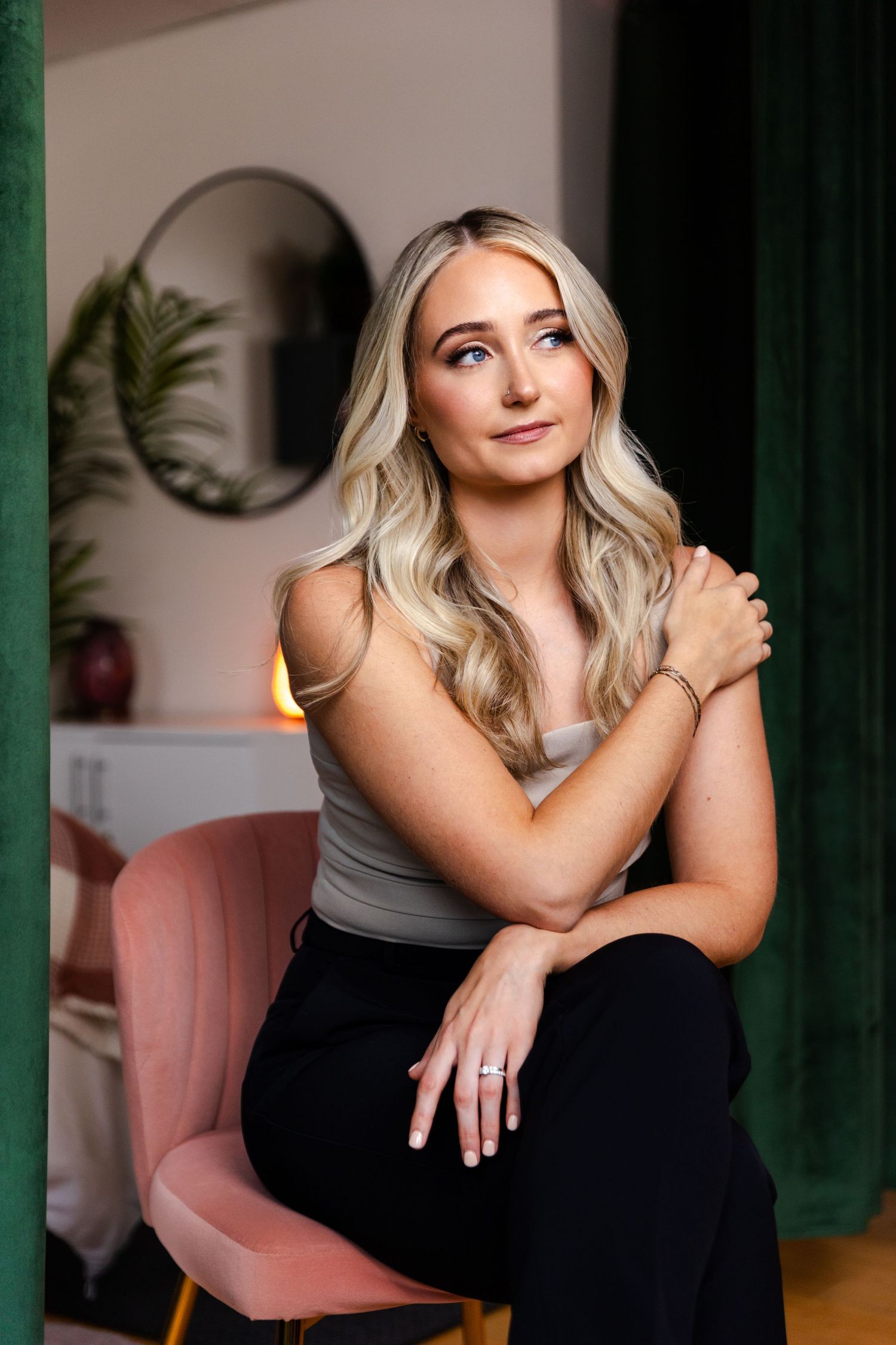 A person posing in a grey tank top and black pants sits on a pink chair, with green curtains and a mirror in background.