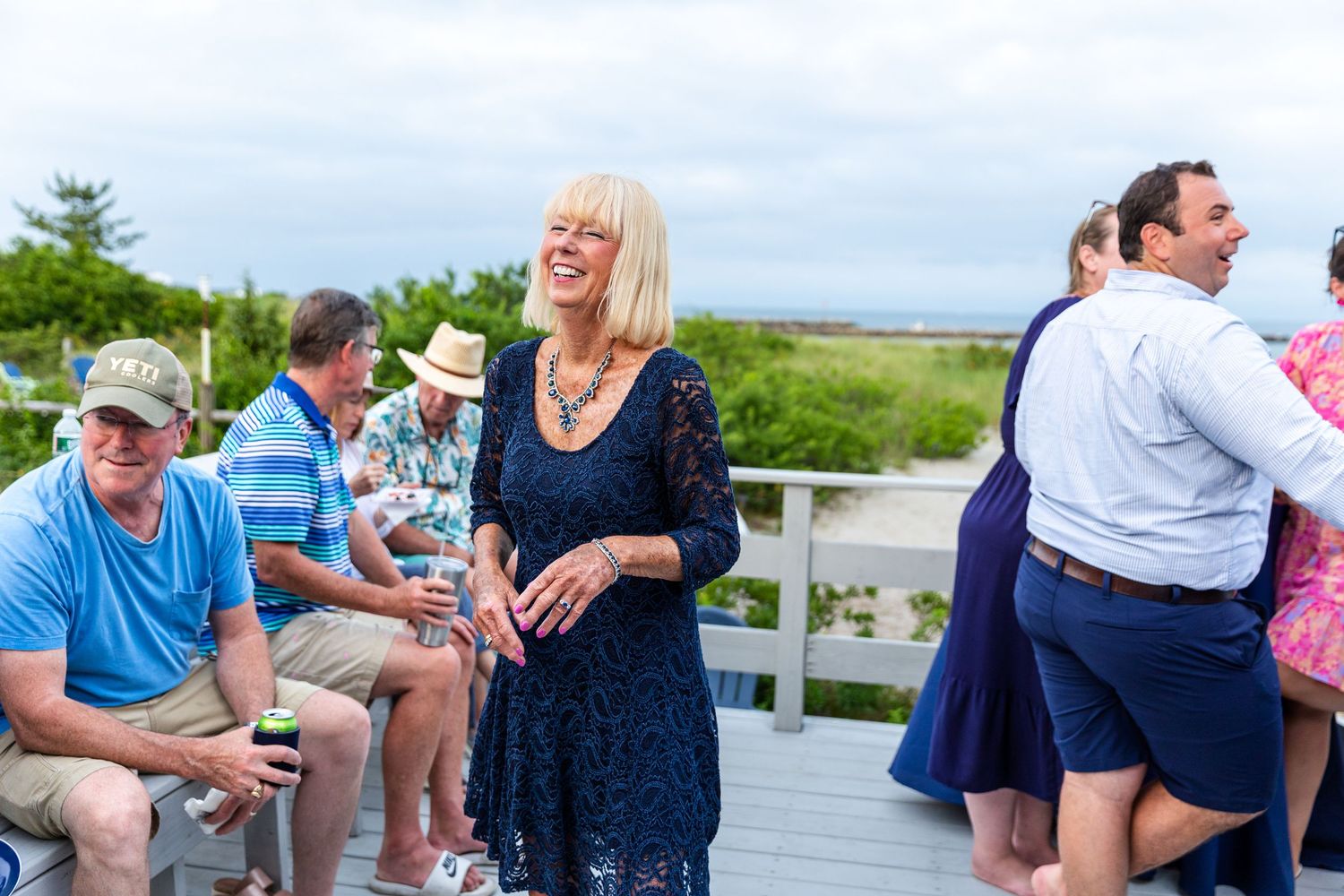 A group of people socializing and laughing together on an oceanside deck during a sunny daytime gathering.