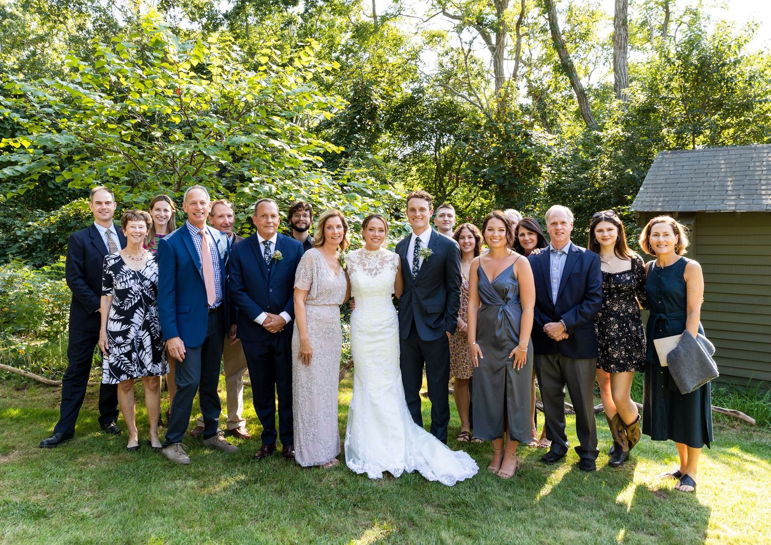 Wedding party group poses together outdoors in formal attire on a sunny garden lawn with trees and greenery.