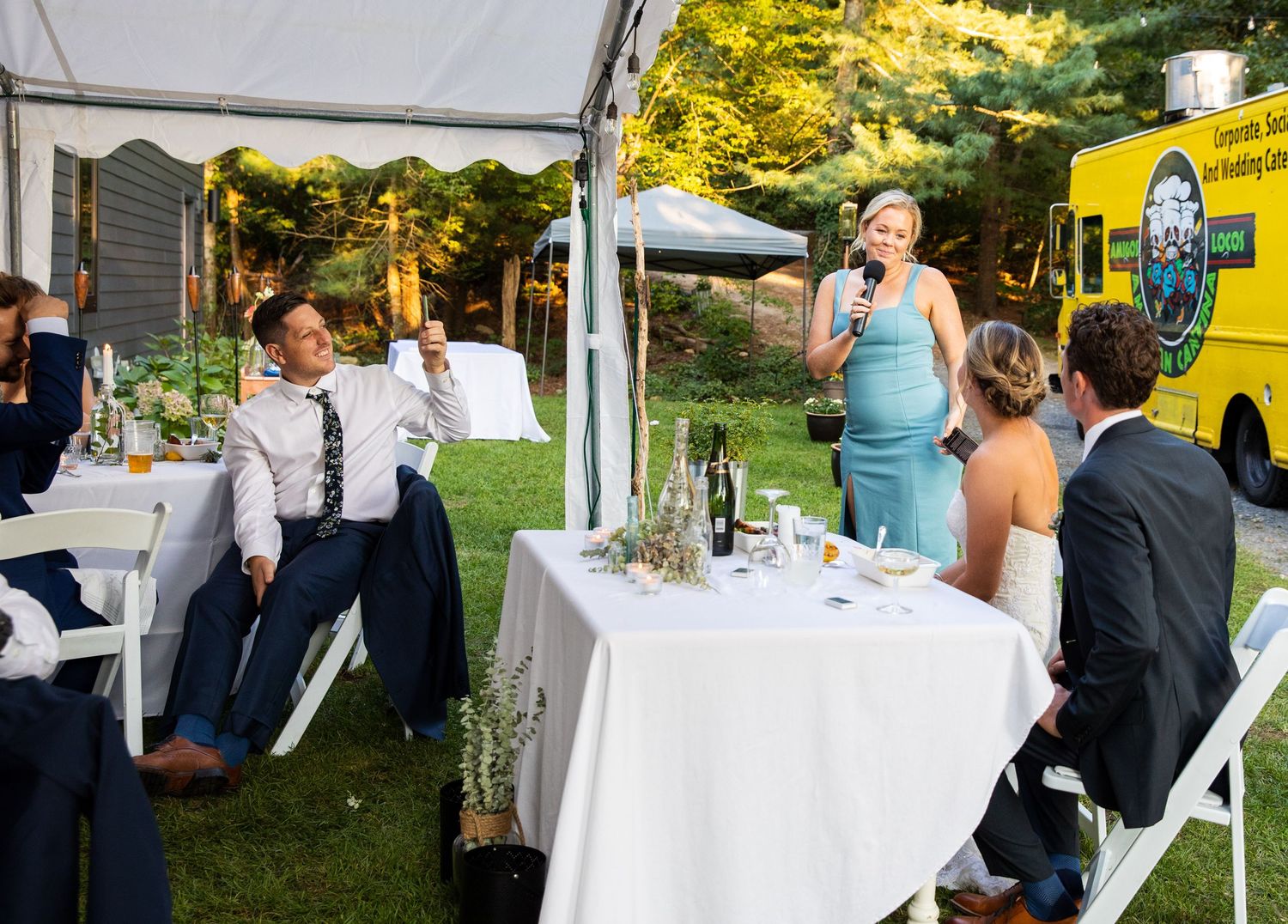 Wedding guests enjoy an outdoor reception under a white tent with a yellow food truck parked nearby on a sunny day.