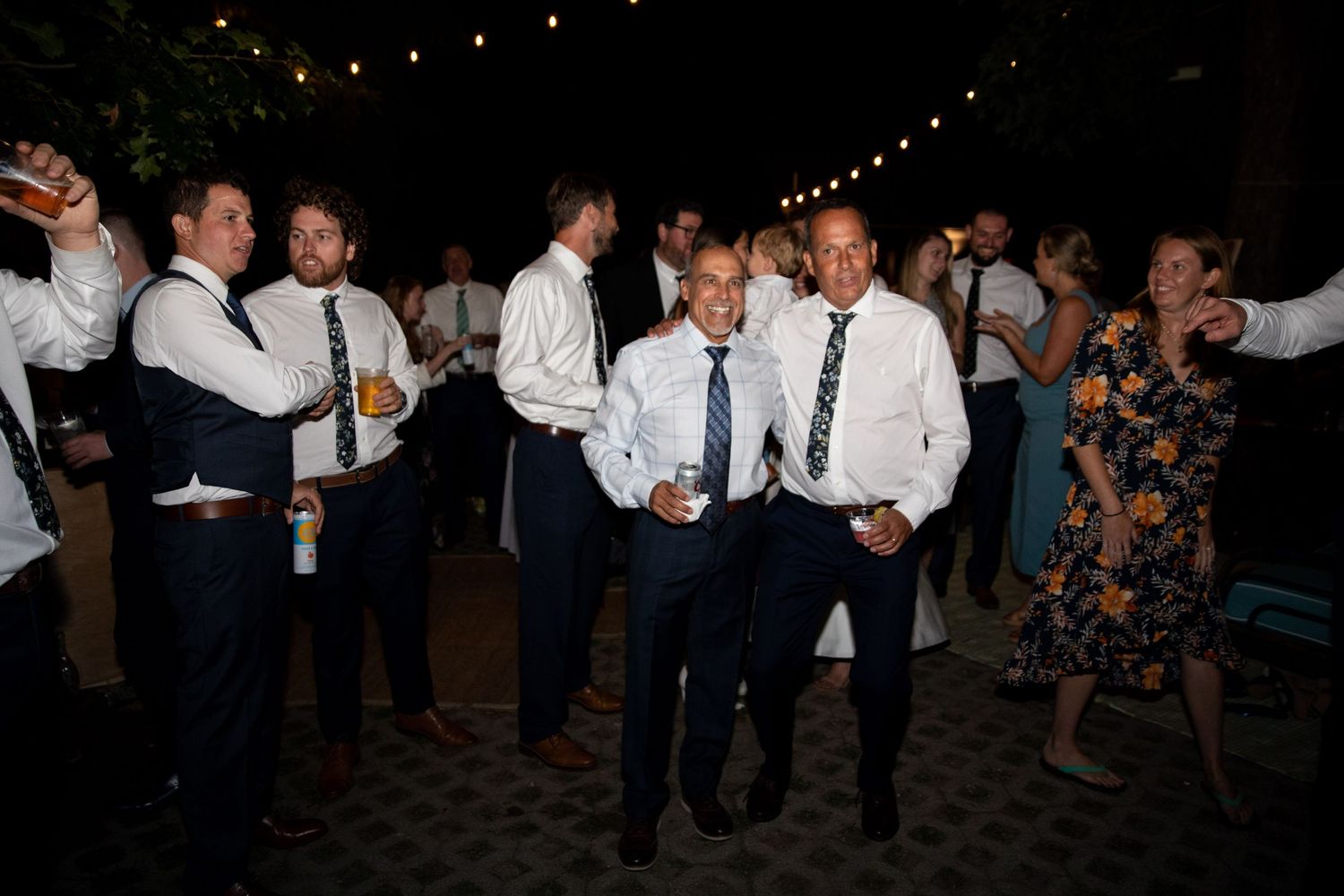 Partygoers in formal attire dancing and mingling under string lights at an outdoor evening celebration.