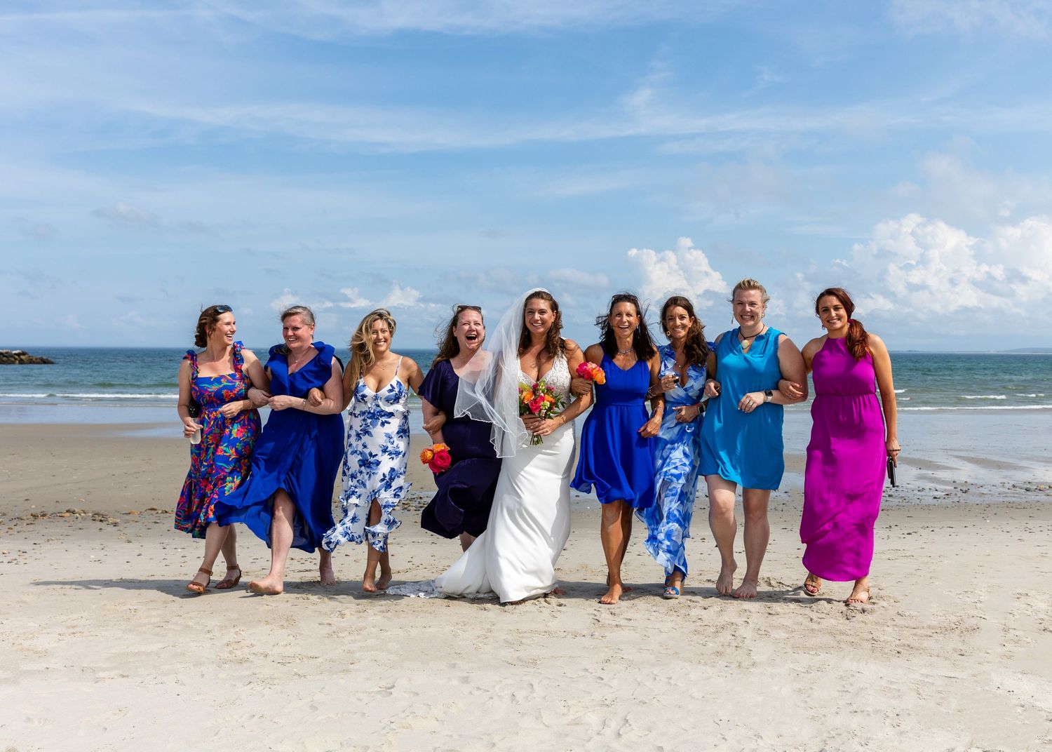 A group of wedding party attendees laugh and walk together on a sunny beach with blue sky and ocean in the background.