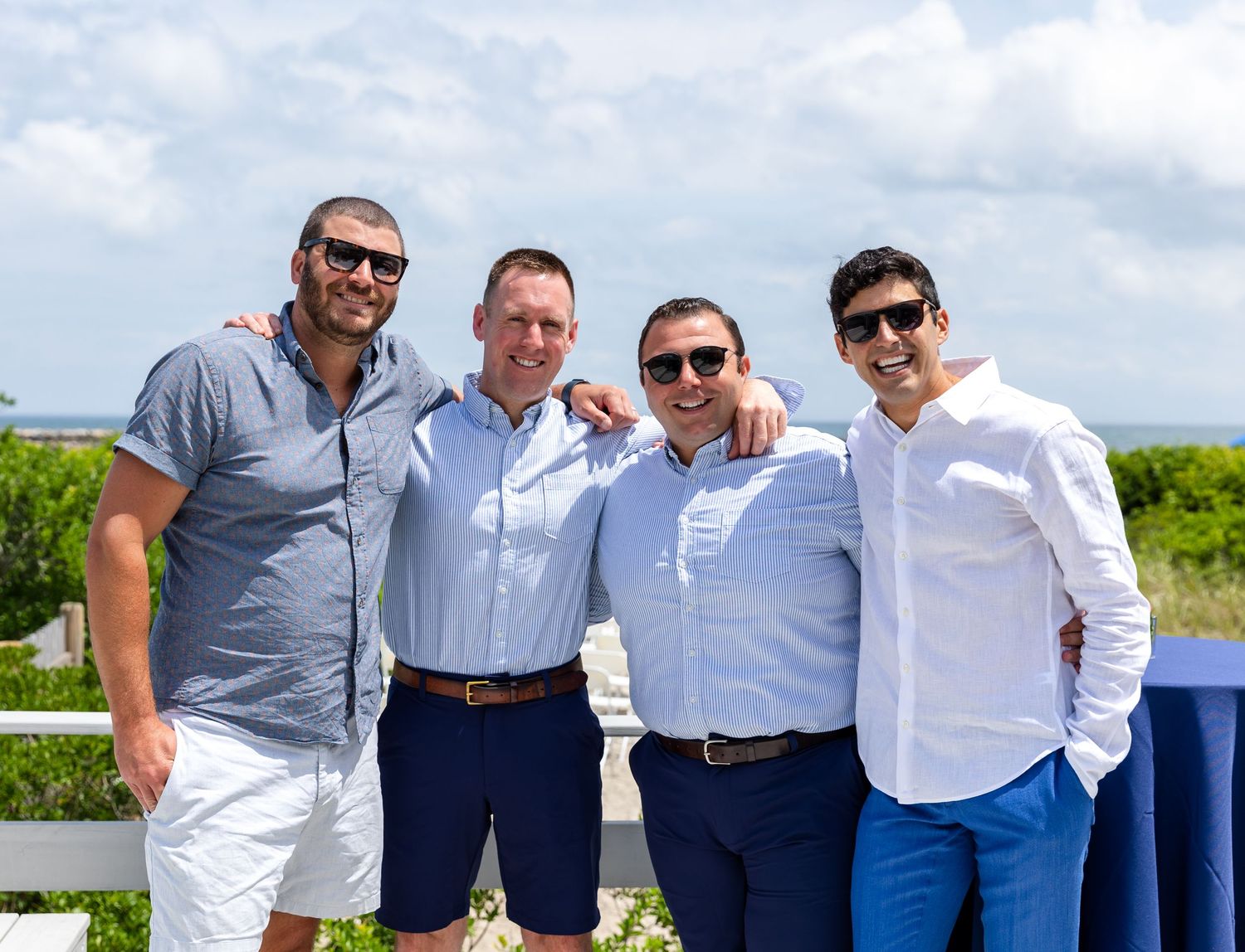 Four confident men wearing sunglasses and button-down shirts pose together outdoors with a scenic view in the background.