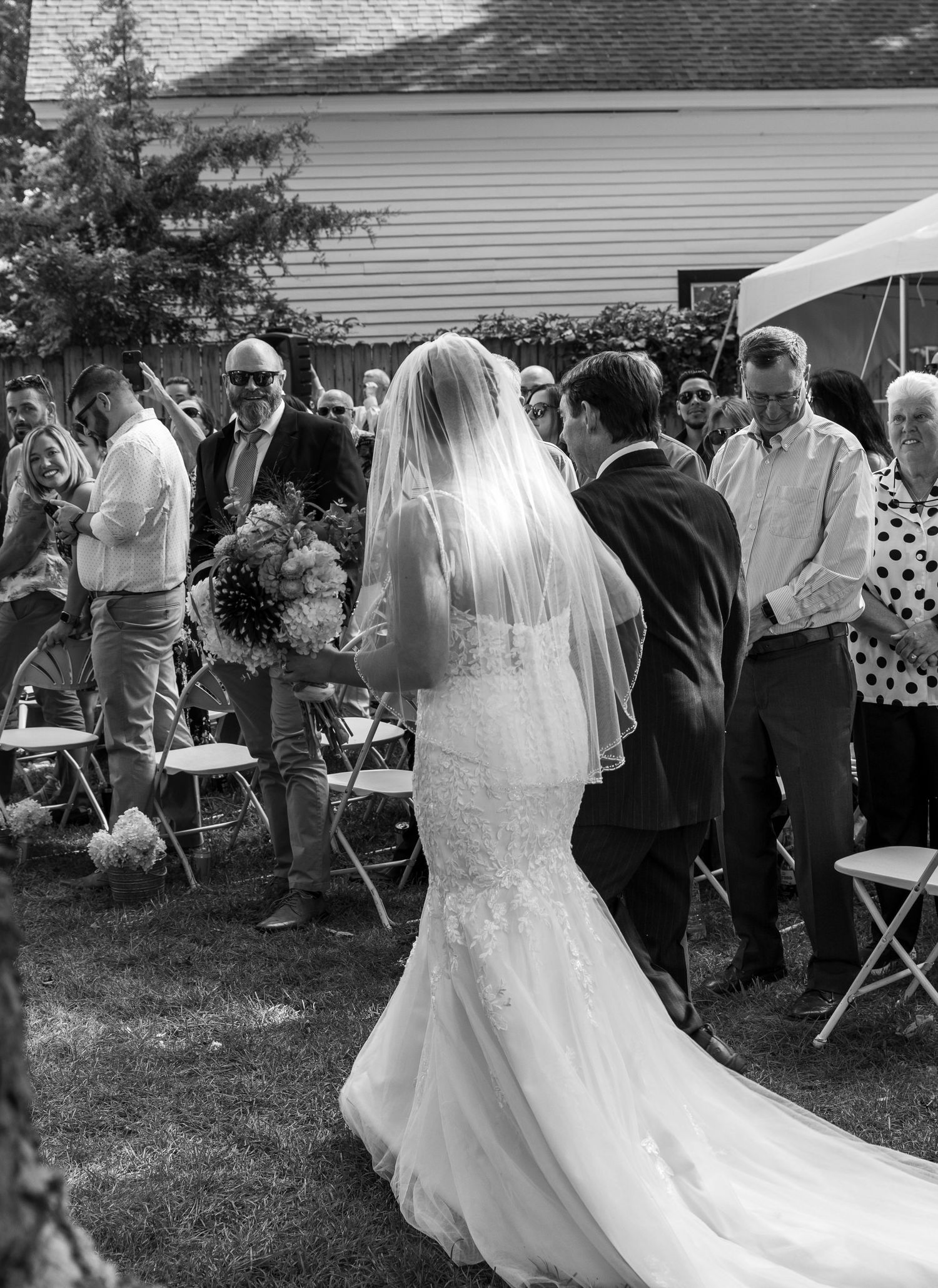 Wedding guests gather outdoors for a backyard ceremony as a bride in a mermaid dress greets attendees.