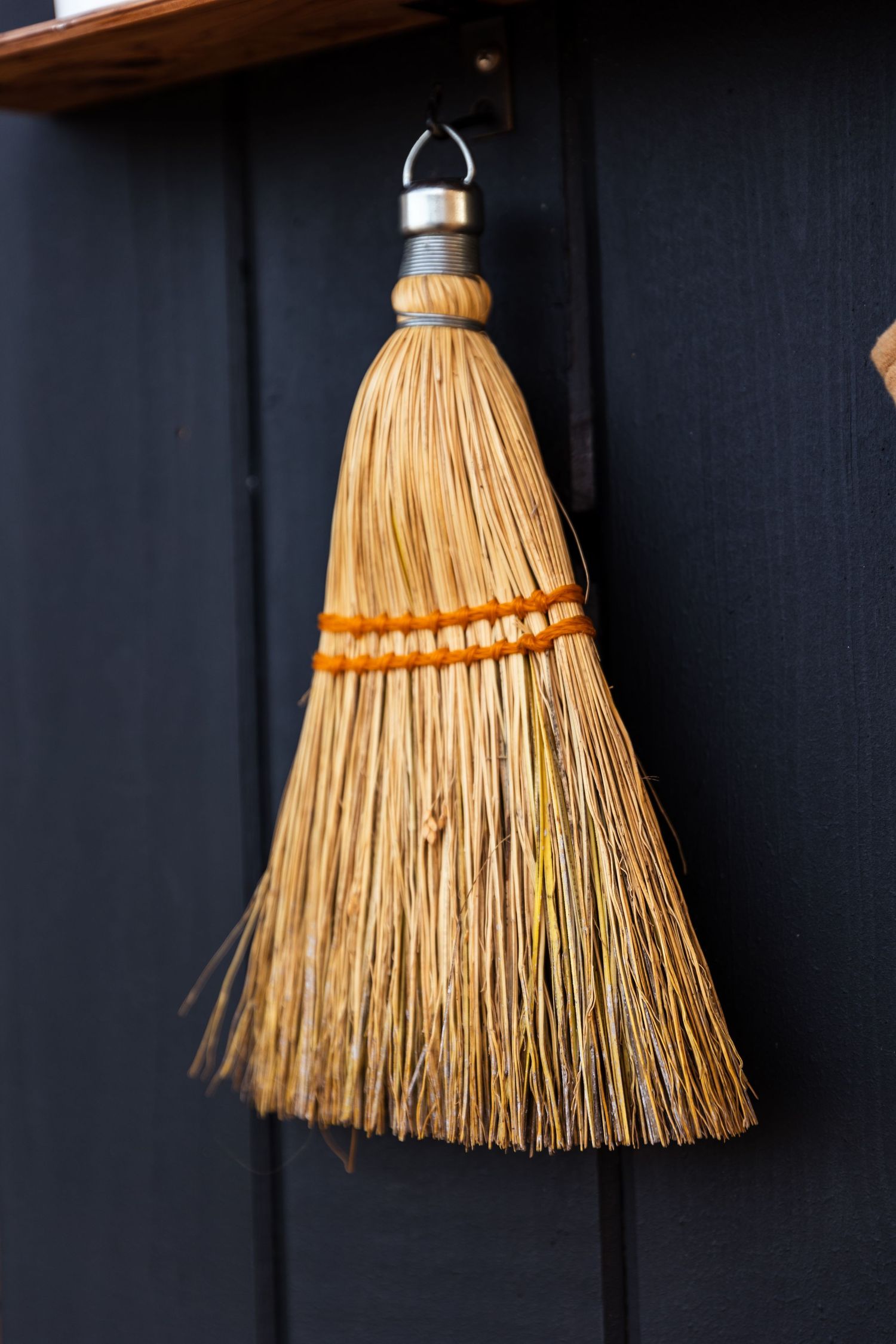 A traditional whisk broom with natural straw bristles and orange decorative band hangs against a dark navy wall.