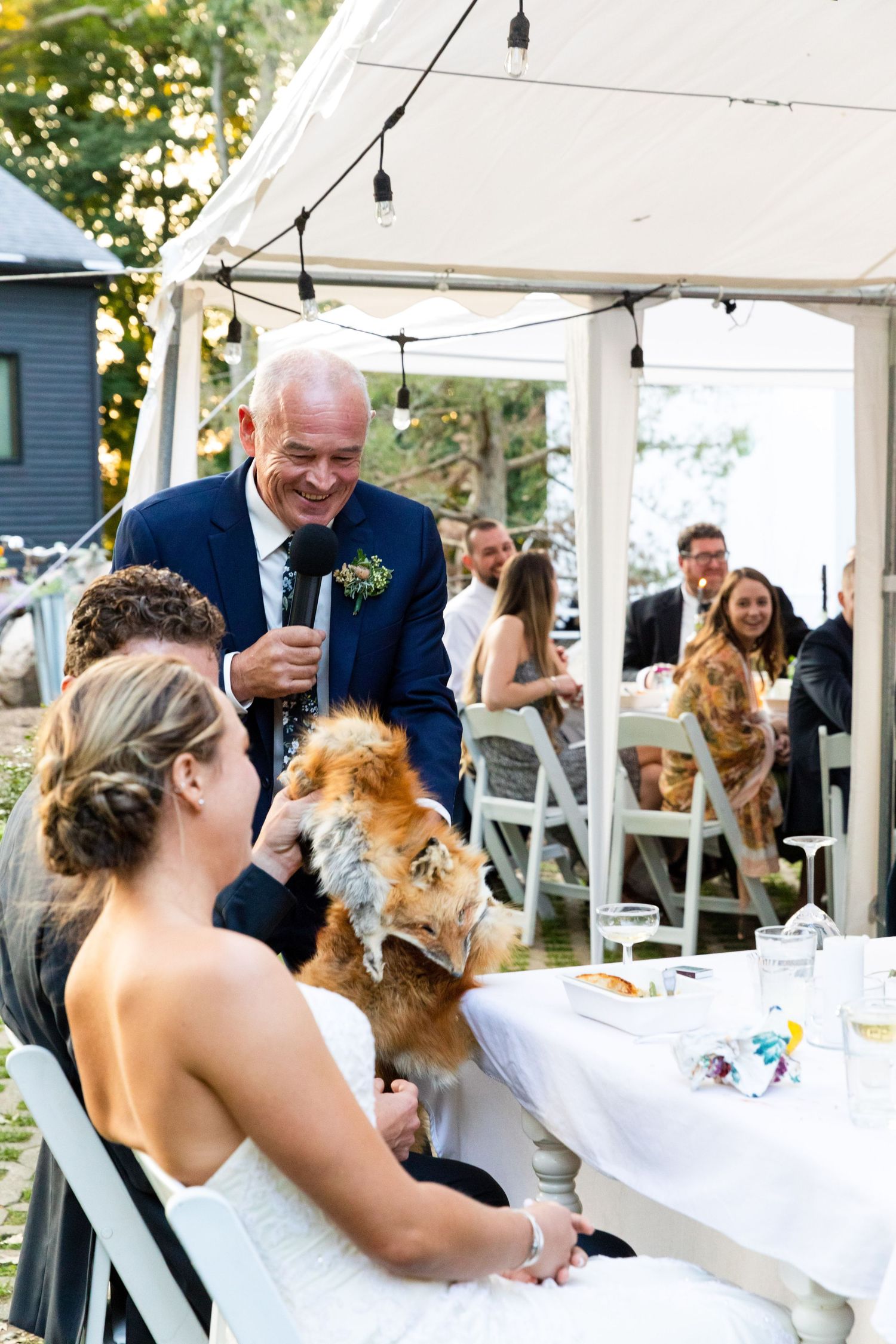 Wedding guests gather under a white tent where two dogs join the celebration at the head table during a reception.