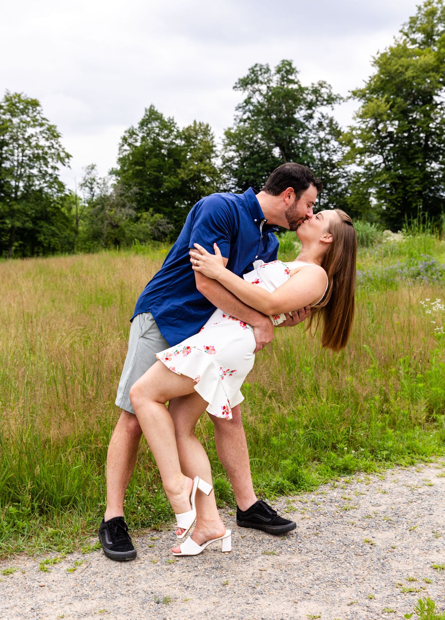 A couple shares a romantic embrace on a dirt path surrounded by tall grass and trees on a summer day.
