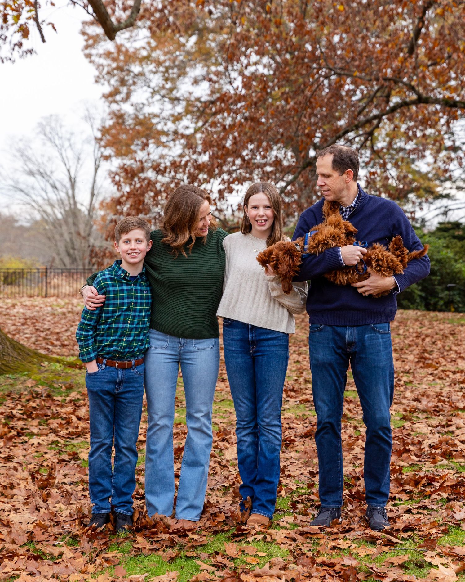 Family stands together outdoors on autumn day surrounded by fallen leaves under a large oak tree with warm brown foliage.