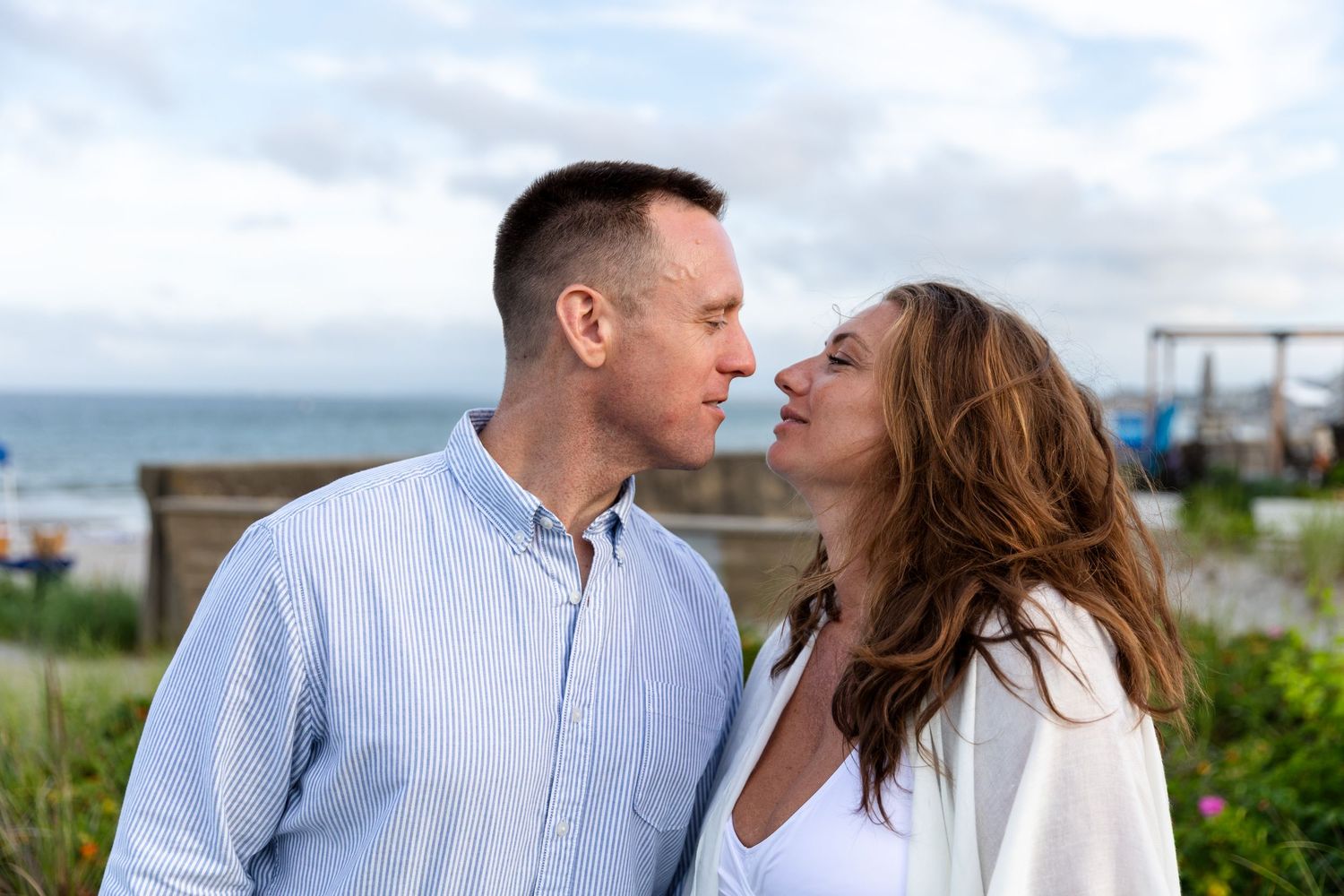 A couple in casual white and blue clothing share an intimate moment together on a coastal overlook by the sea.