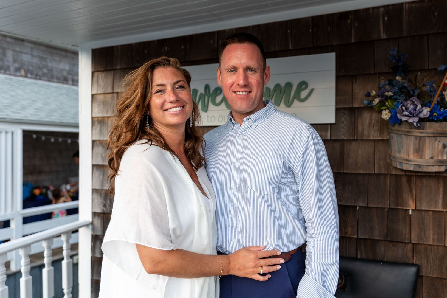 A couple in casual white shirts pose together on a wooden porch with a 'welcome' sign behind them.
