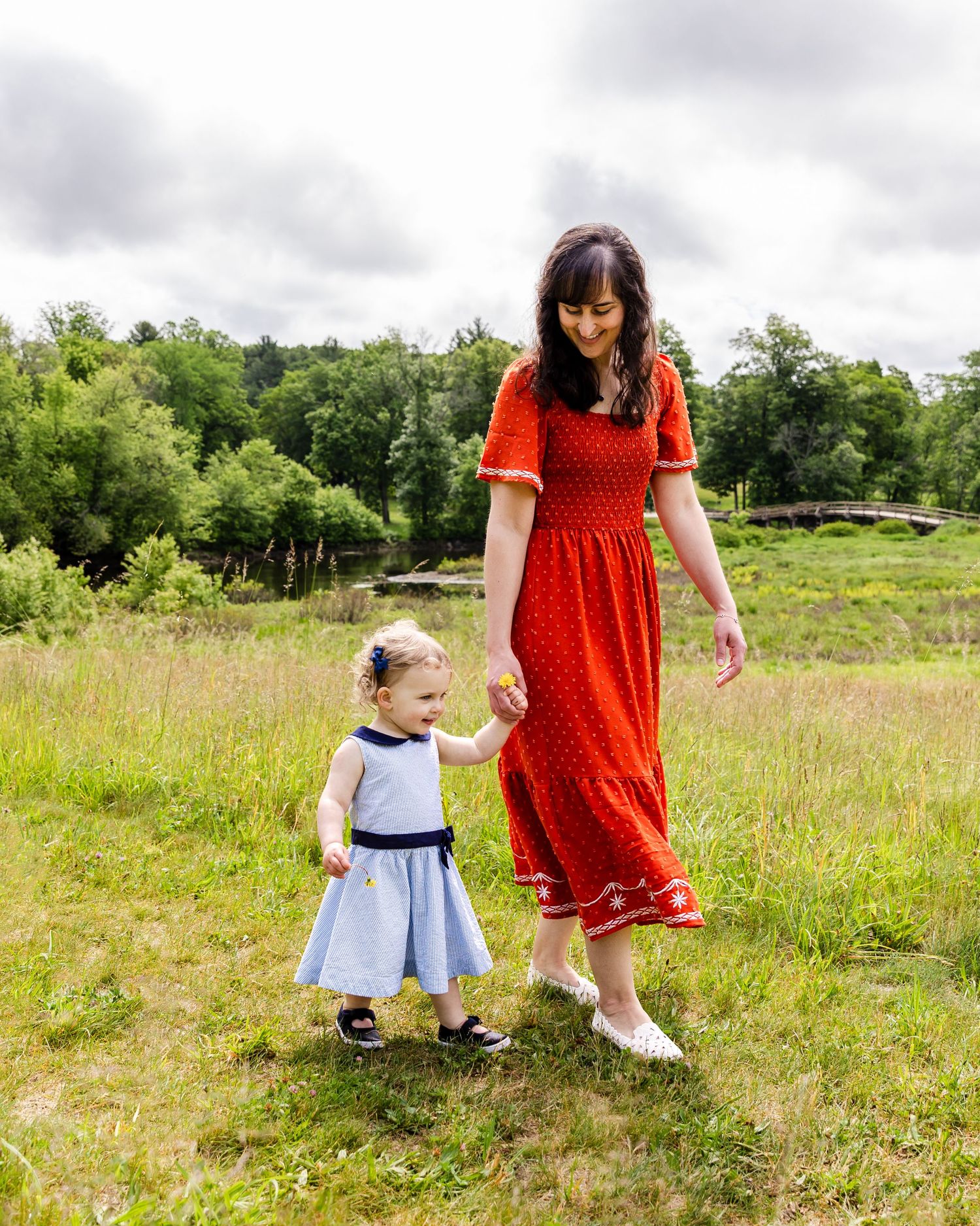 Adult in red polka dot dress and child in blue dress hold hands while walking together in a grassy field with trees.